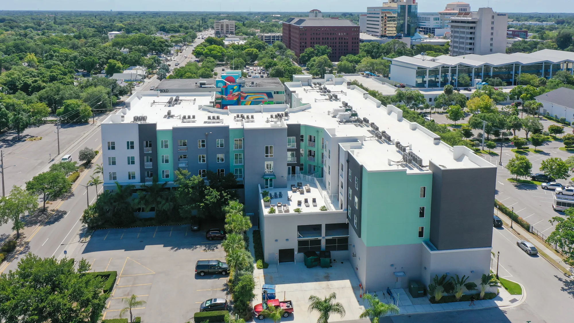An aerial view of a modern multi-storey building featuring a green and gray facade, surrounded by lush greenery and commercial properties. The image showcases the building's terrace area with seating, as well as nearby streets and parking areas.