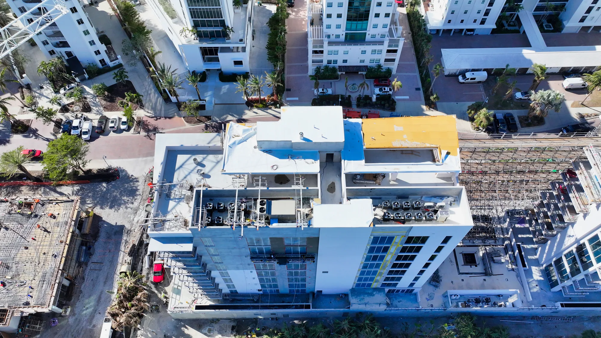 The image shows an aerial view of a multi-story building under construction, with visible rooftop structures and ongoing work on adjacent buildings. Surrounding the site, there are parked vehicles and landscaped areas, indicating a developing urban environment.