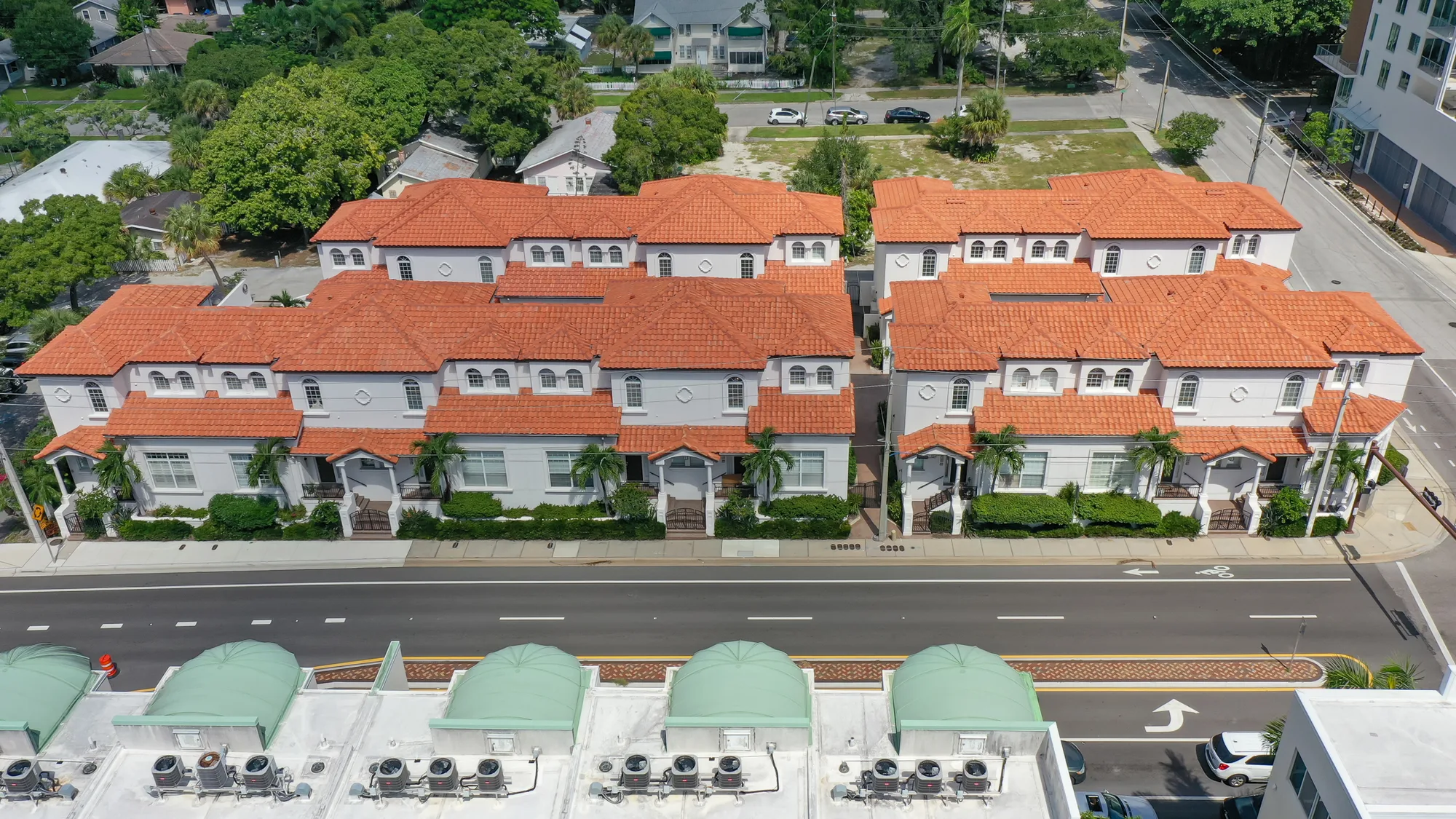 An aerial view of a residential building complex featuring multiple units with red tile roofs and white exteriors. The complex is set alongside a road lined with greenery and additional houses in the background.