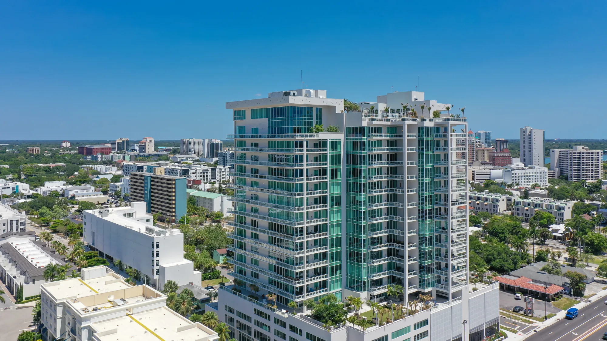 Aerial view of a contemporary high-rise building featuring glass balconies and rooftop greenery, surrounded by various urban structures and greenery in a city landscape.