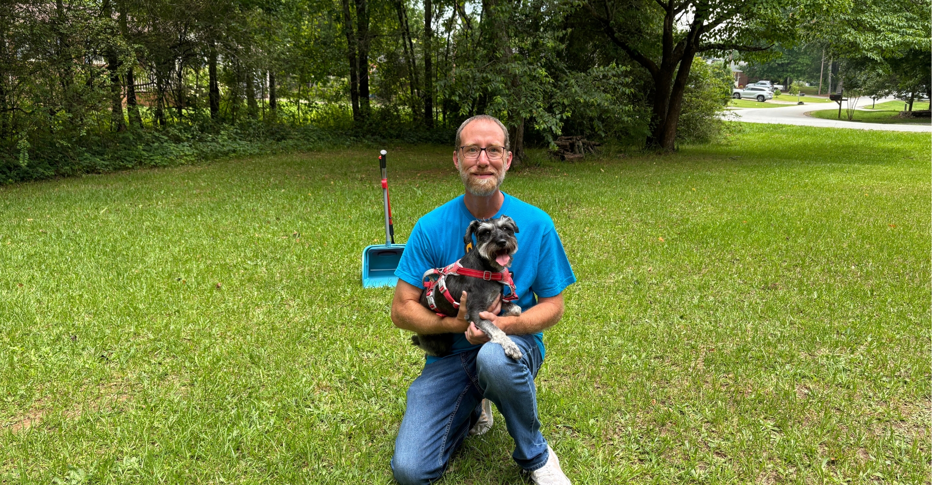 Man kneeling in a grassy area holding a small dog, with a dog waste scoop visible in the background
