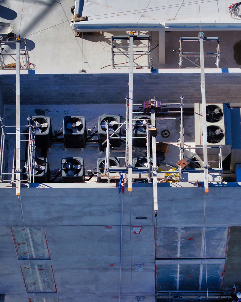An aerial view of a construction site showing an array of rooftop air conditioning units and scaffolding. The image captures unfinished concrete walls and construction materials, indicating ongoing building work.