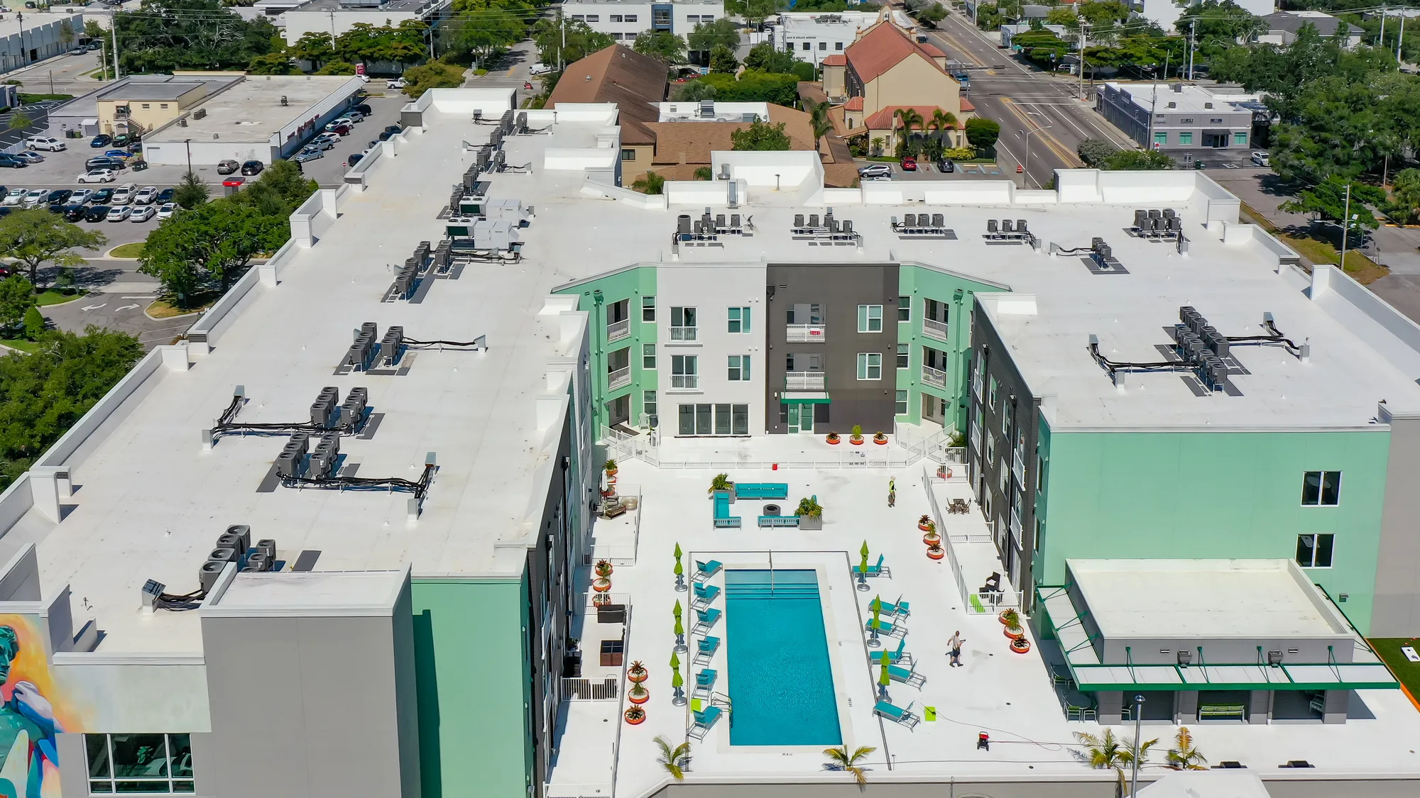 The image shows aerial view of a modern multi-story apartment complex featuring a central swimming pool surrounded by lounge chairs and green umbrellas. The building has a mix of green and grey exterior, and the area is landscaped with plants and seating arrangements.