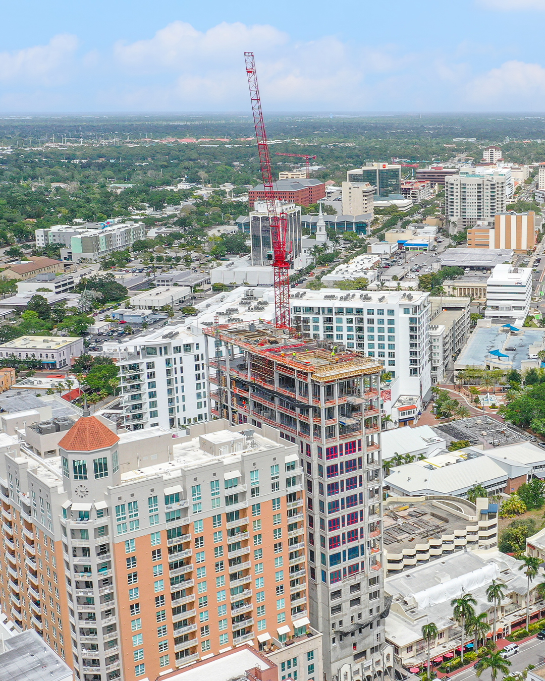 The image shows a high-rise building under construction in an urban area, featuring a prominent red crane on top. Nearby structures include other residential and commercial buildings, with greenery visible in the background.