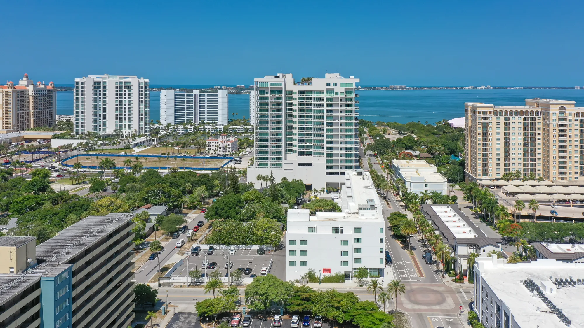 This aerial image showcases a coastal city with high-rise buildings, palm trees, and ocean views. The foreground features a mix of modern and older architecture, with a vibrant urban landscape under a clear blue sky.