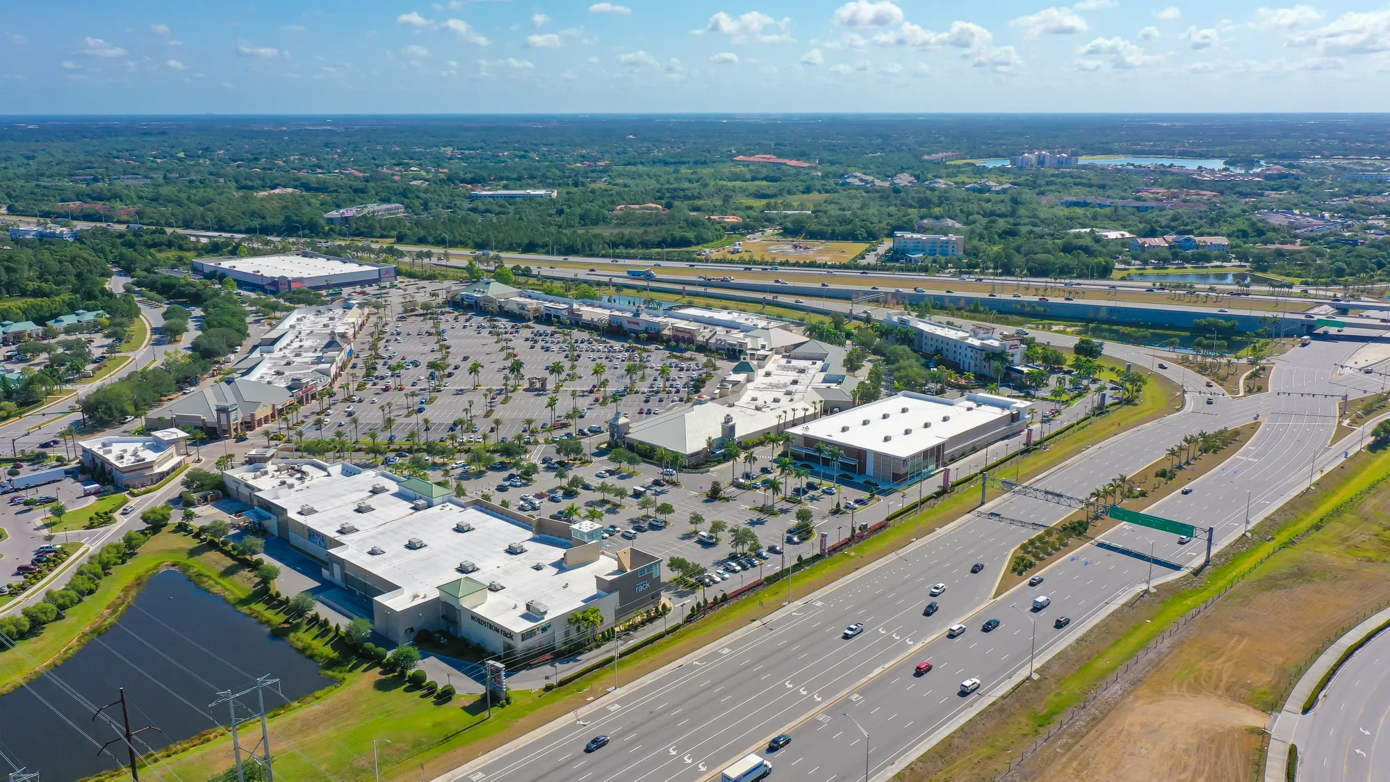 This aerial image showcases a large shopping complex featuring extensive parking and nearby highway. Lush green areas and additional commercial buildings are visible in the background, indicating a developed urban landscape.