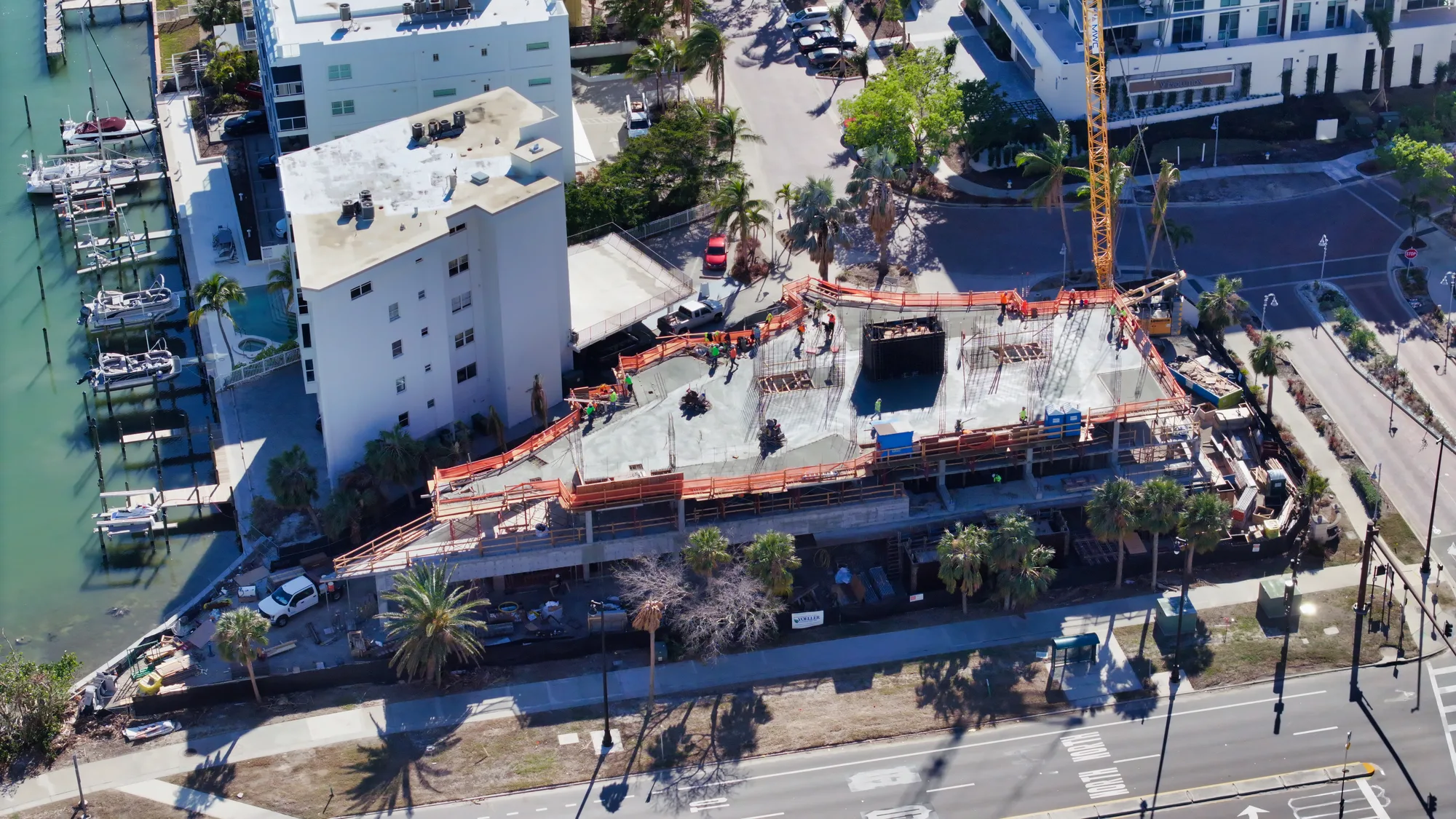 An aerial view of an active construction site featuring a concrete foundation being laid, surrounded by palm trees and nearby buildings. Construction workers and equipment are visible, indicating ongoing development in the area.