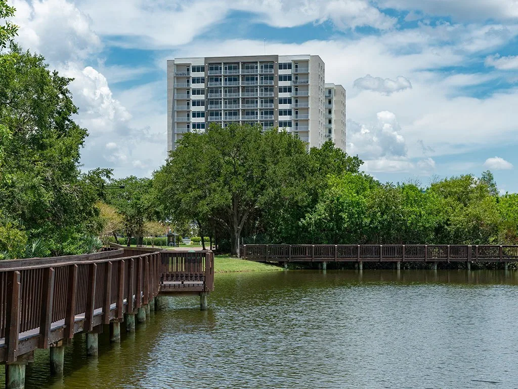 The image depicts a tranquil waterfront scene featuring a wooden boardwalk winding through greenery, with a tall residential building visible in the background against a partly cloudy sky.