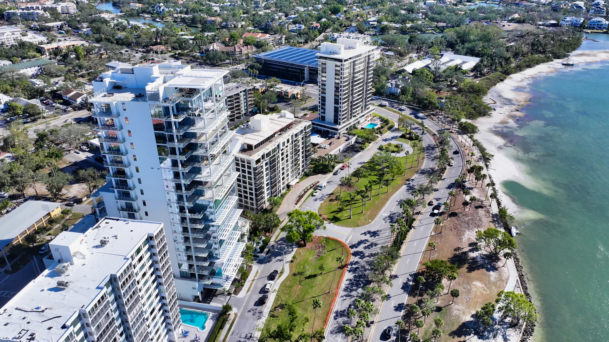 An aerial view showcases a coastal apartment complex featuring multiple high-rise buildings near a shoreline. The image captures landscaped areas, swimming pools, and a roadway lining the waterfront, with greenery and the ocean visible in the background.