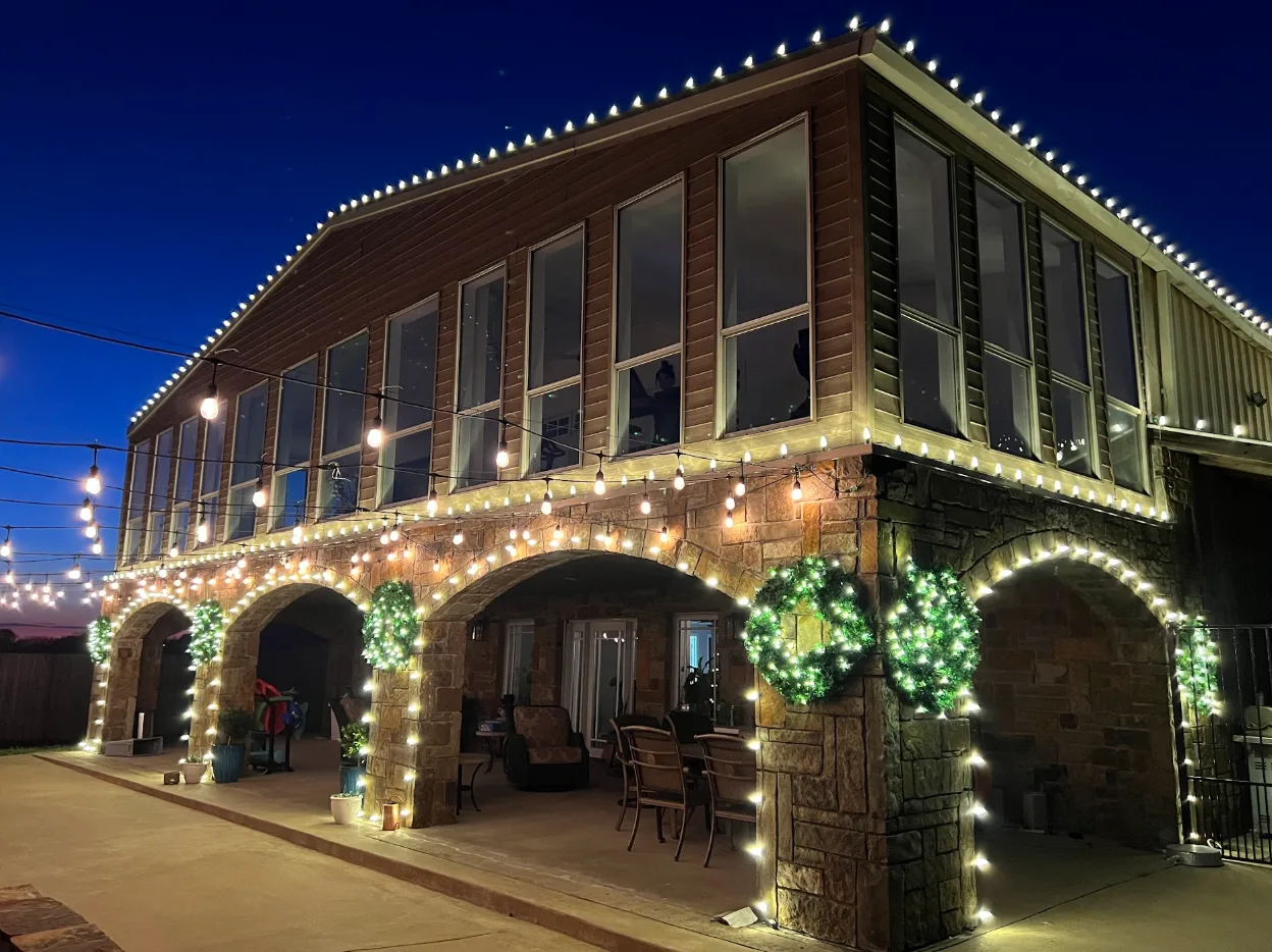 Large stone and timber venue photographed at dusk, with a lit roofline, wreaths hung between stone arches, garland wrapped around the columns and bistro string lights strung across the patio.