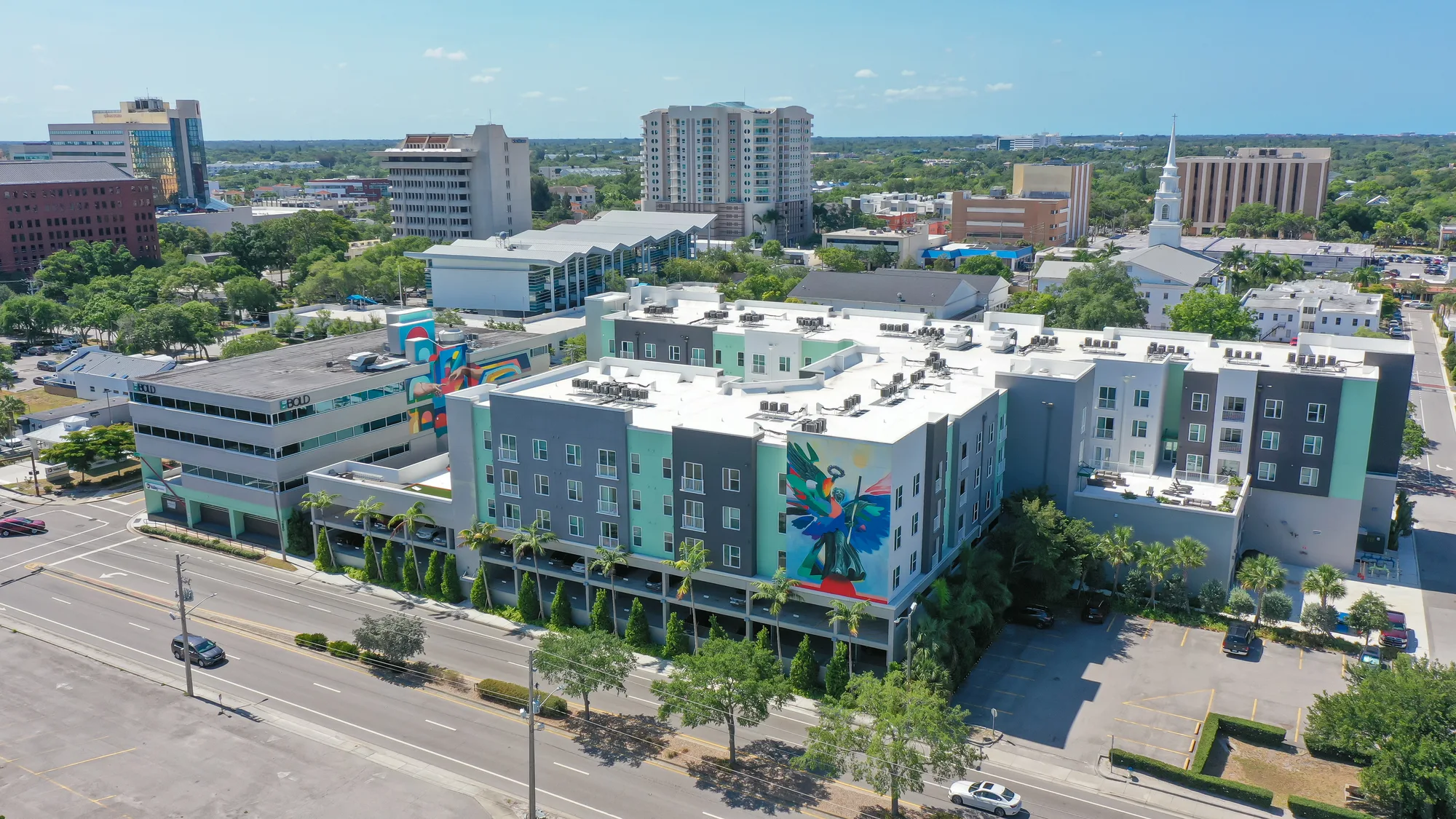 An aerial view of a modern apartment complex featuring vibrant murals on its exterior. Surrounding buildings and a busy street can be seen in the background, highlighting the urban setting.