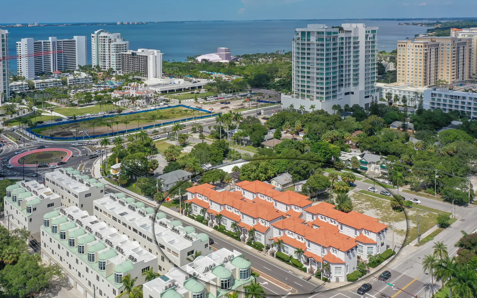 This aerial image showcases a coastal development area featuring low-rise buildings with red tile roofs, surrounded by lush greenery and a waterfront. The background reveals modern high-rise buildings and urban infrastructure near a body of water, highlighting a mix of residential and commercial properties.
