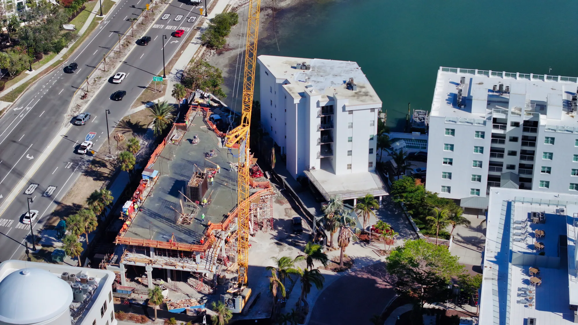 An aerial view of an active construction site featuring a large crane and ongoing building work. Nearby, residential buildings are visible along a roadway adjacent to a body of water.