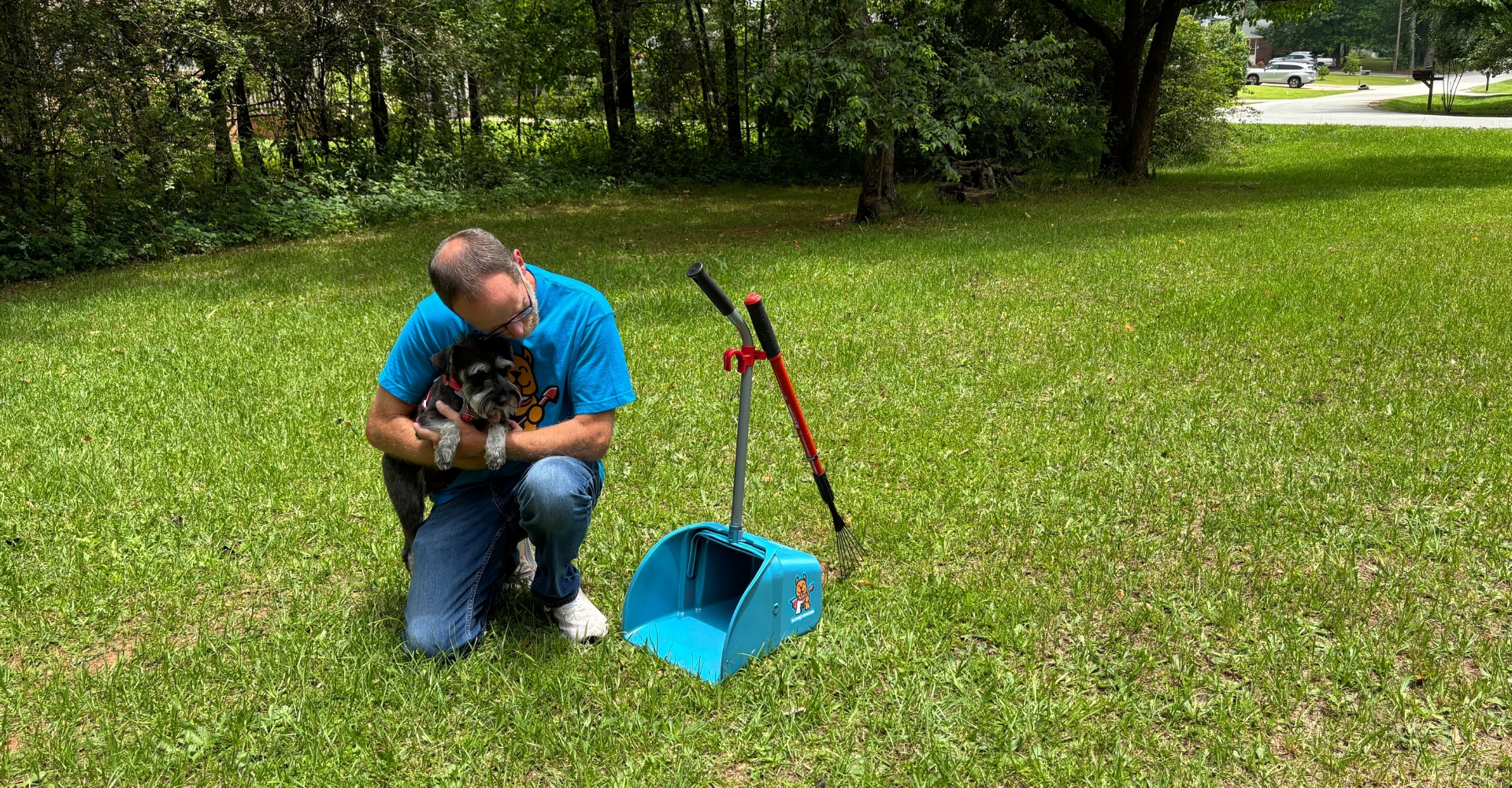 Man kneeling in yard cuddling a small dog next to a pet waste scooper