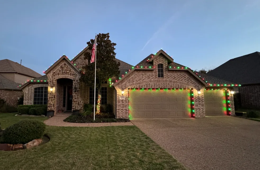 Brick home photographed at dusk with alternating red and green bulbs along the rooflines and both garage doors outlined.