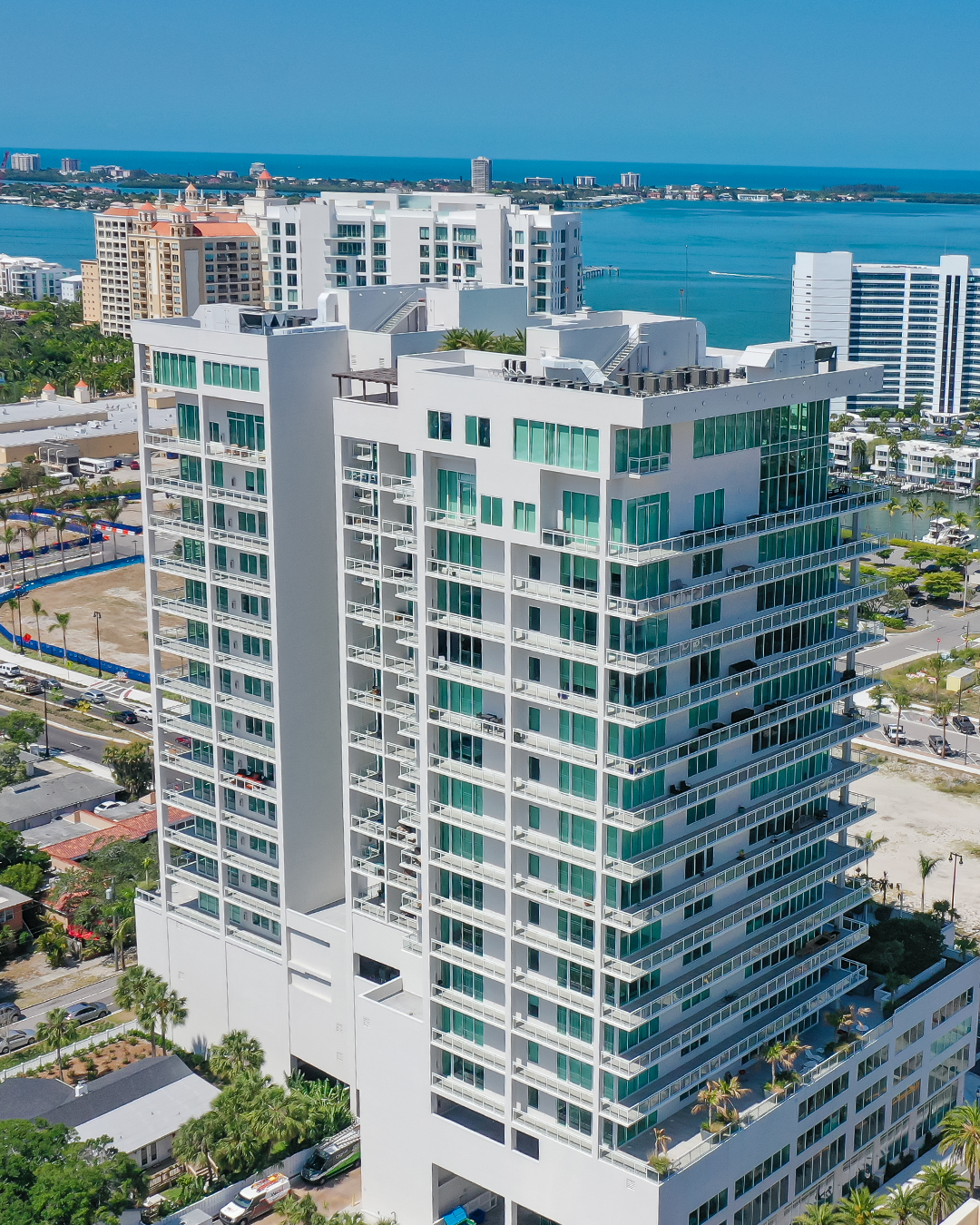 Aerial view of a modern high-rise apartment building featuring large glass windows and multiple balconies, with the ocean and urban landscape visible in the background. Lush greenery and nearby low-rise structures can also be seen at the base of the building.