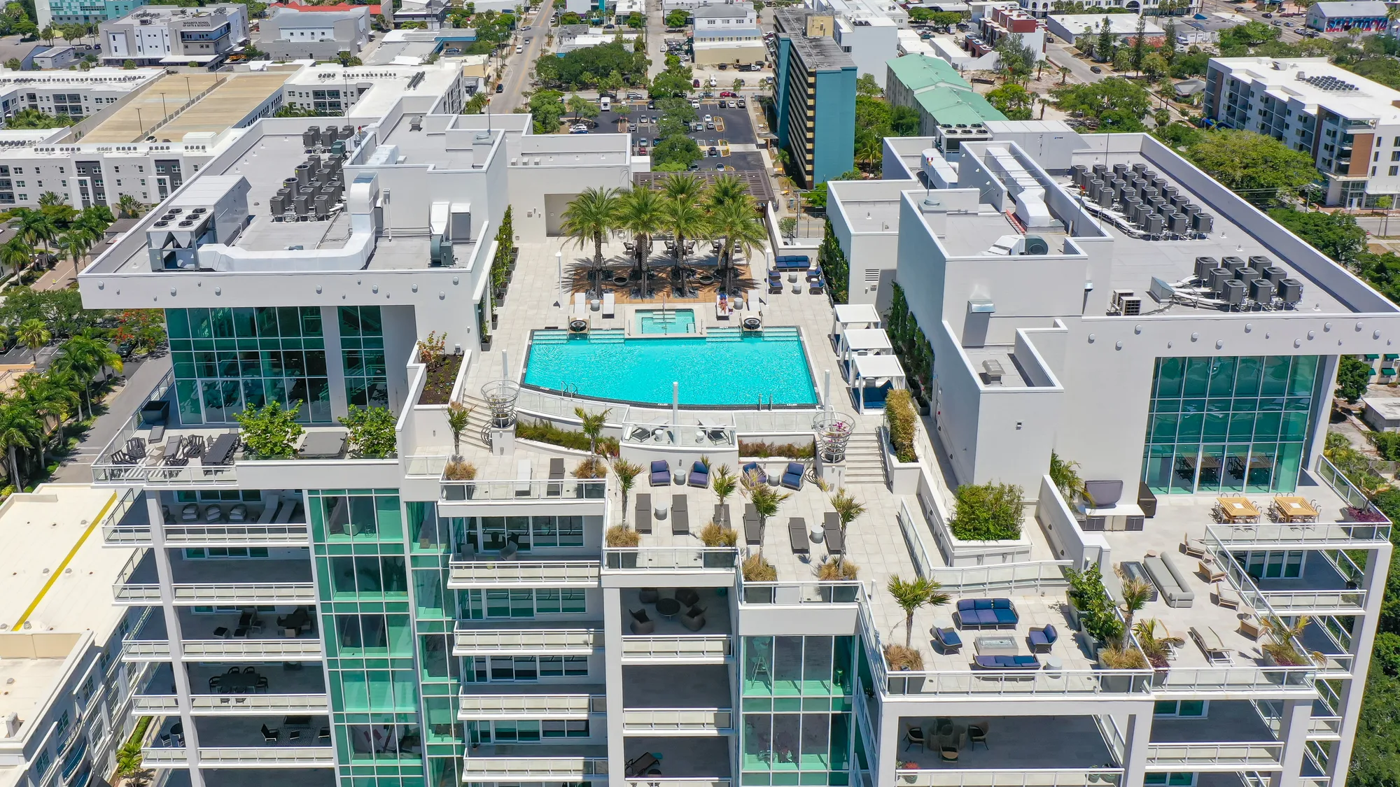 An aerial view of a contemporary residential complex featuring a large, clear swimming pool surrounded by lounge chairs and palm trees. The building has expansive glass windows and roofs, showcasing an upscale living environment.