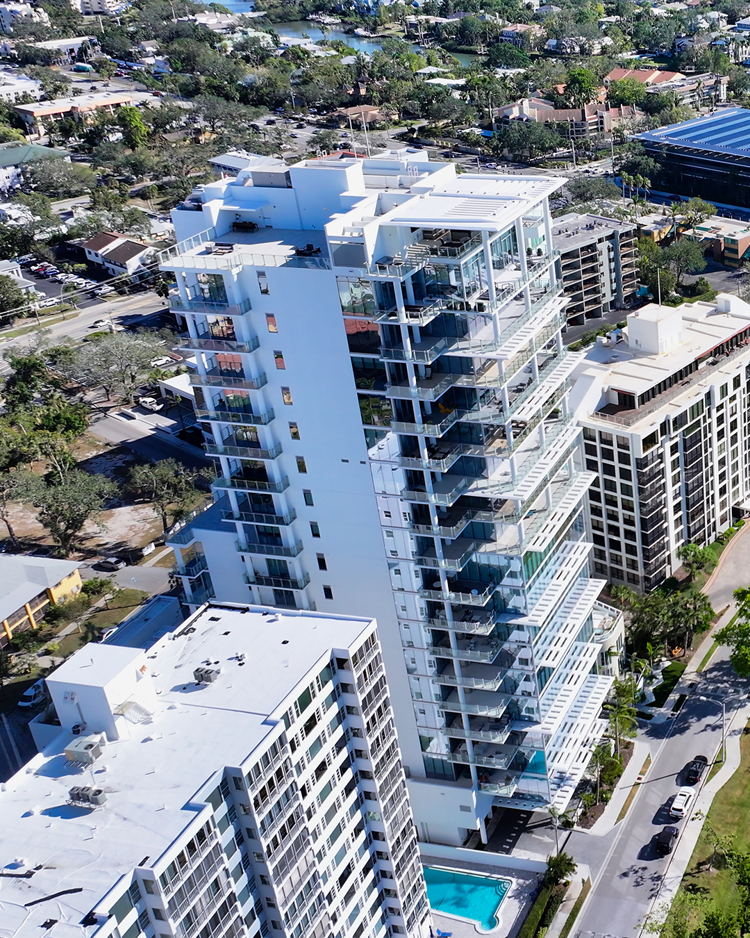 An aerial view of a modern high-rise building featuring glass balconies and a rooftop terrace, surrounded by residential areas and a swimming pool at its base. The surrounding landscape includes a mix of trees and streets, typical of a suburban setting.