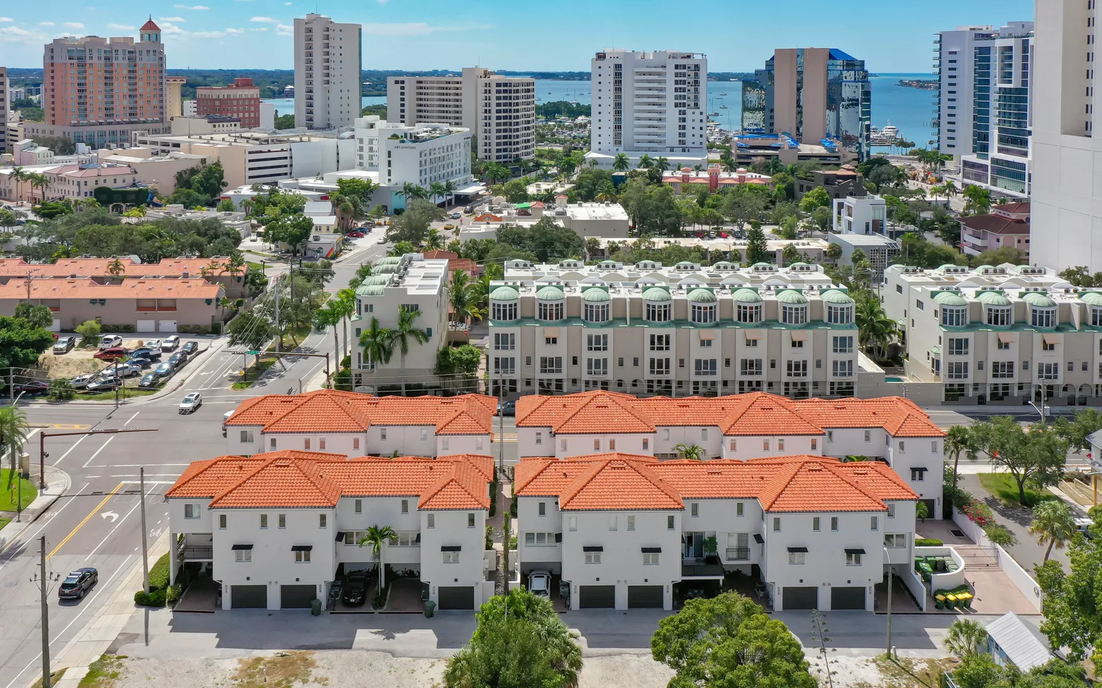 An aerial view of a residential area featuring houses with red tiled roofs, surrounded by modern high-rise buildings and a waterfront in the background. The image captures a mix of urban development, including residential and commercial spaces.