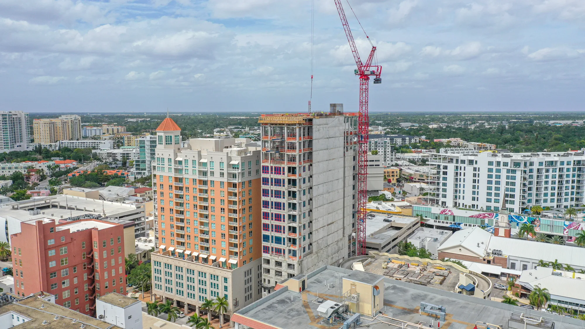 An aerial view of a construction site featuring a partially completed high-rise building alongside other buildings in a bustling urban area. A red crane is actively involved in the construction, with a mix of residential and commercial structures visible in the backdrop.