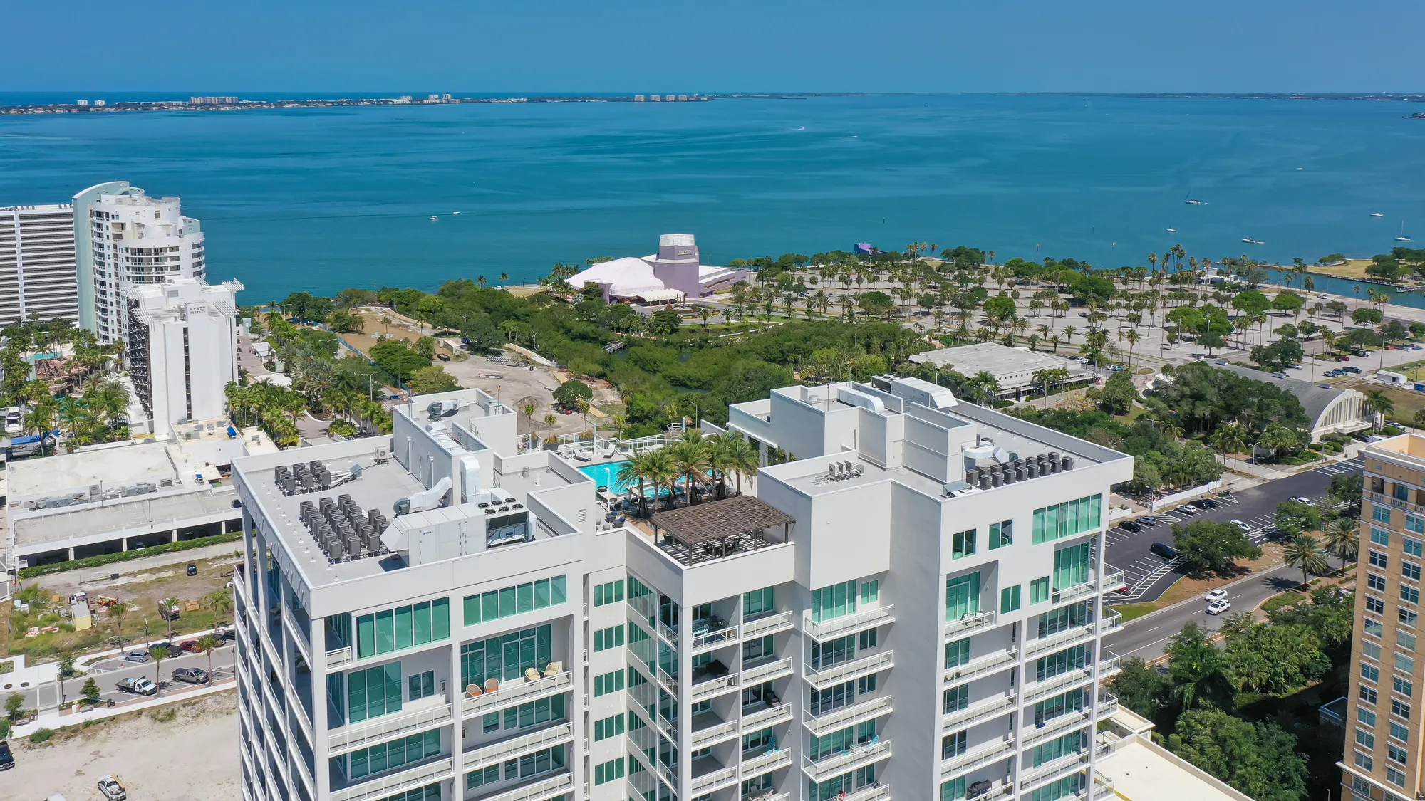 The image showcases aerial view of modern high-rise buildings adjacent to a coastal area, featuring greenery and open spaces. The blue waters of the bay are visible in the background, with a prominent pink building and palm trees in the foreground.