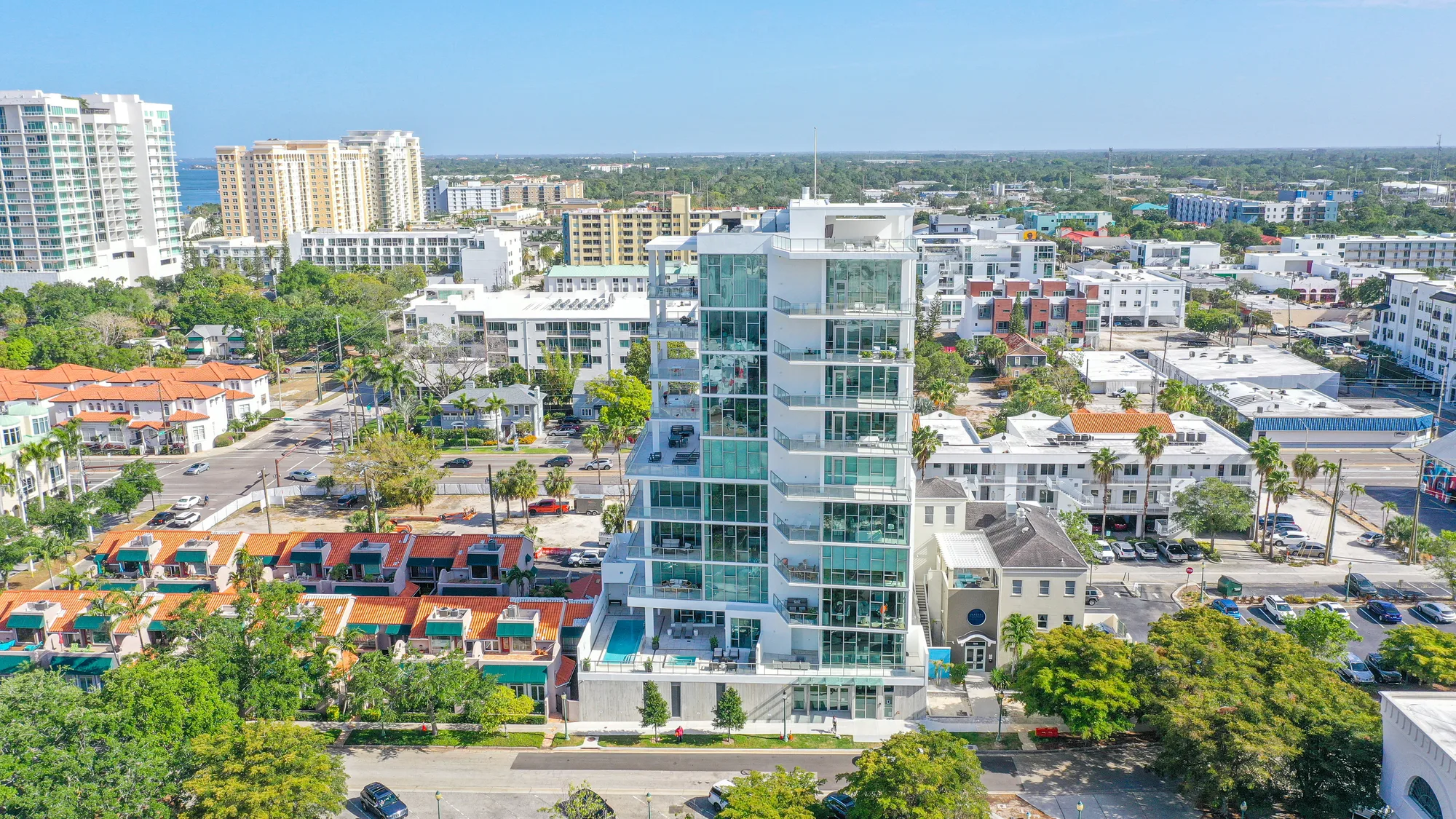 An aerial view of a modern high-rise building surrounded by a mix of residential and commercial structures, featuring glass balconies and a rooftop pool. The vibrant city landscape includes low-rise homes and palm trees, under a clear blue sky.