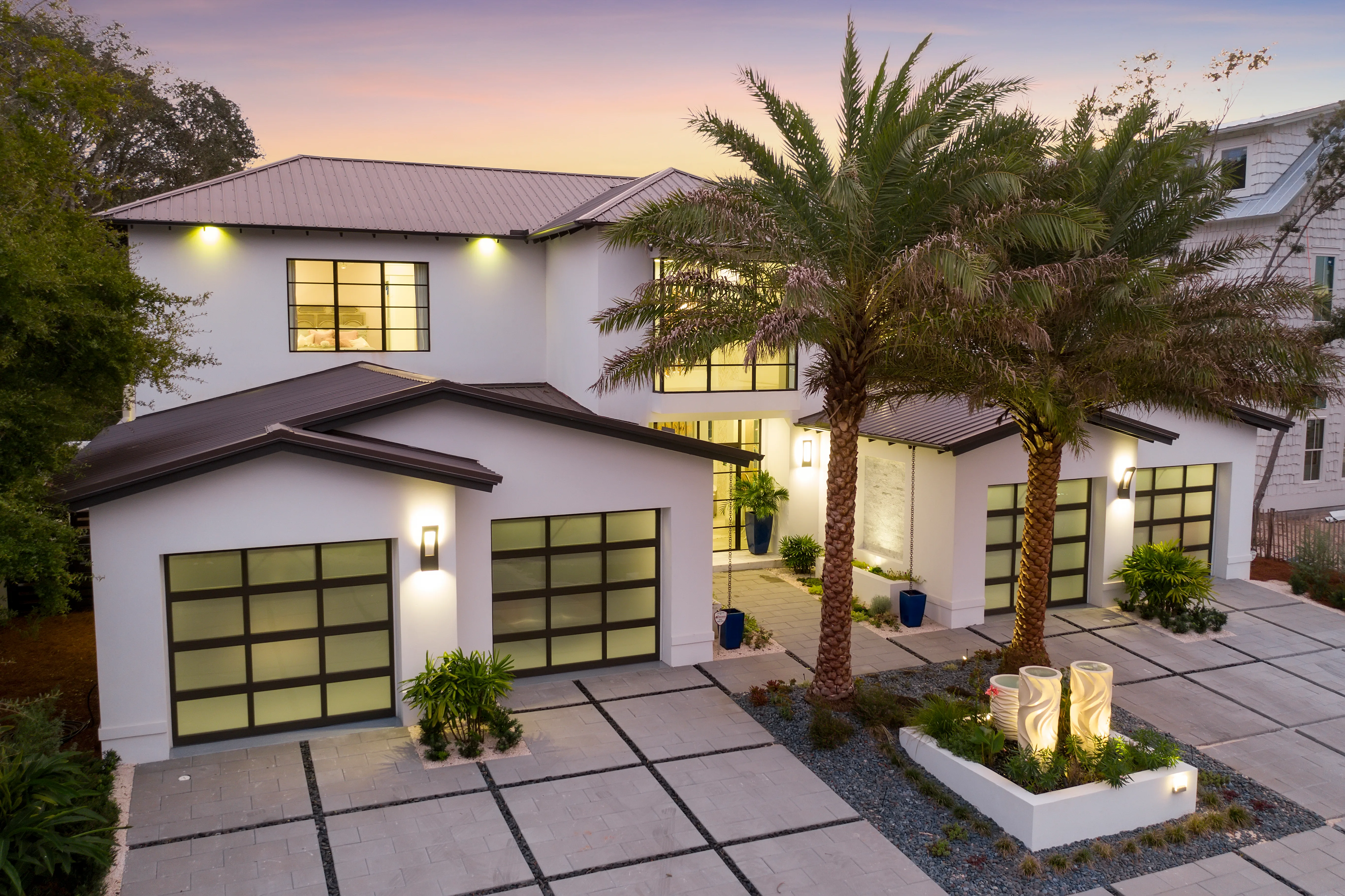 A finished home at dusk with lit glass garage doors and palm trees in the foreground.