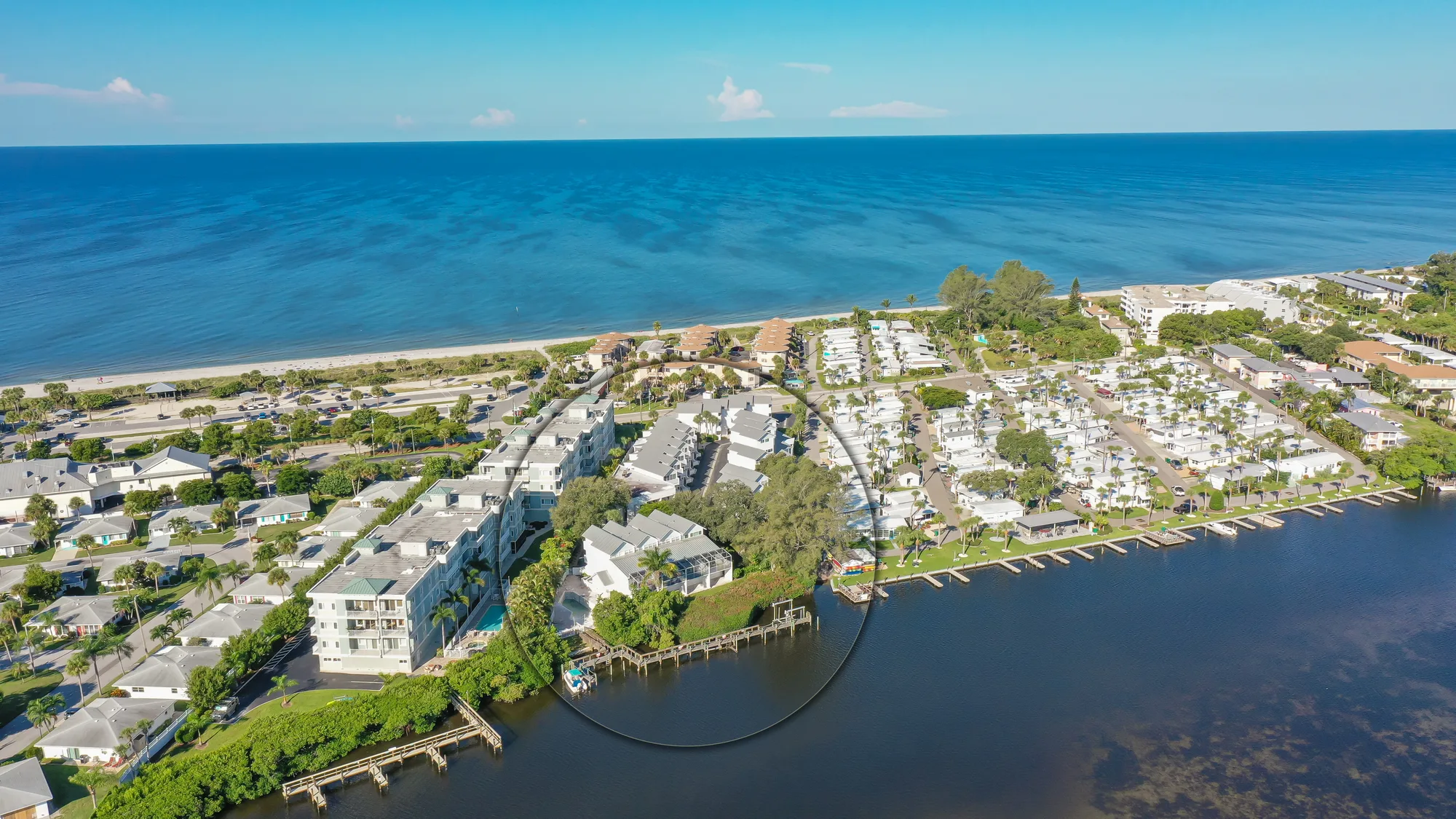 This aerial image showcases a coastal community along a beach, featuring a mix of residential buildings and a marina beside a calm blue ocean. The layout includes various homes, palm trees, and parking areas adjacent to the shoreline.