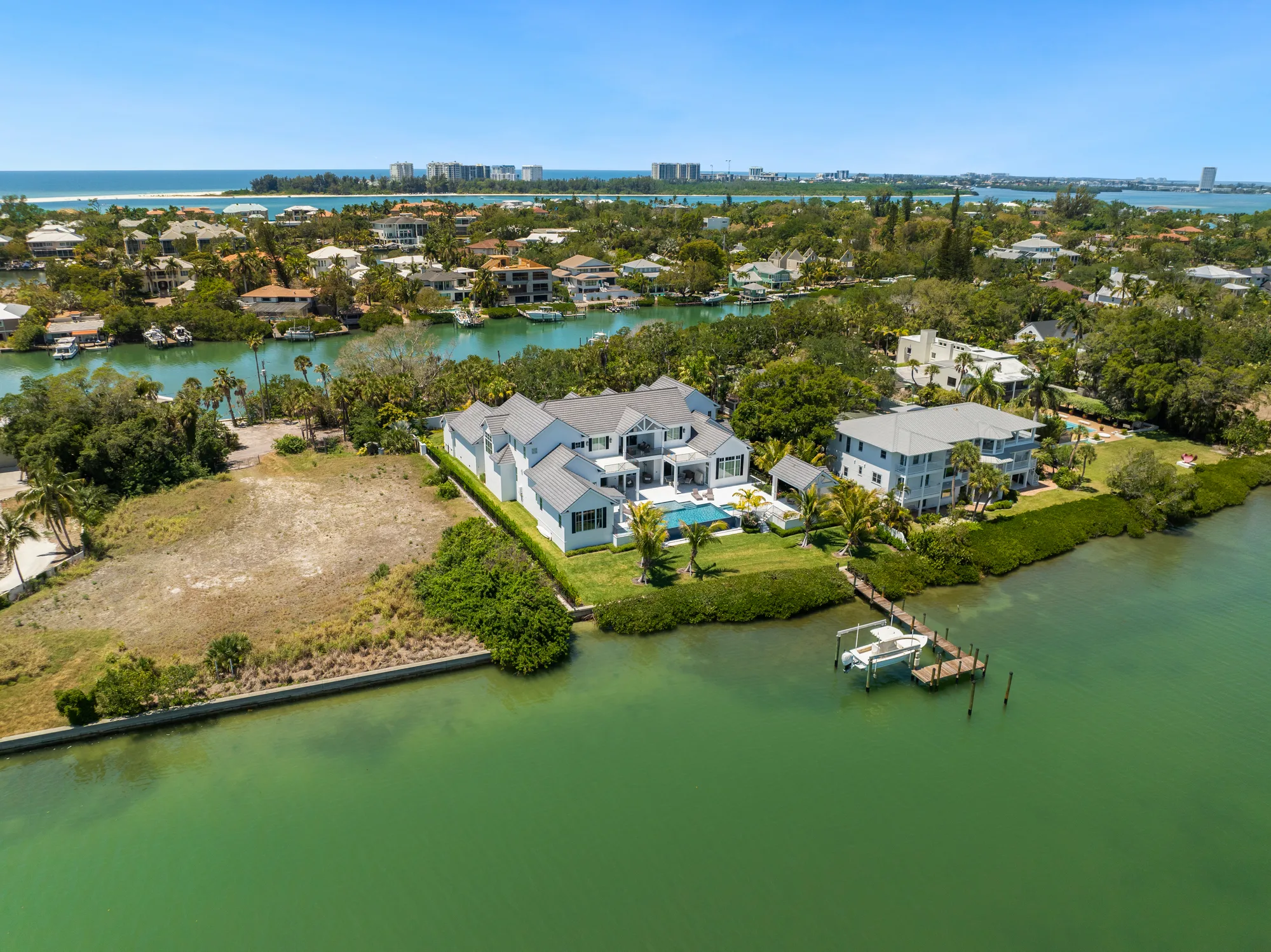This aerial image captures a residential area with waterfront homes, lush greenery, and clear blue waters. Notable features include a spacious house with a pool and a dock, set against a backdrop of other homes and a distant view of the coastline.