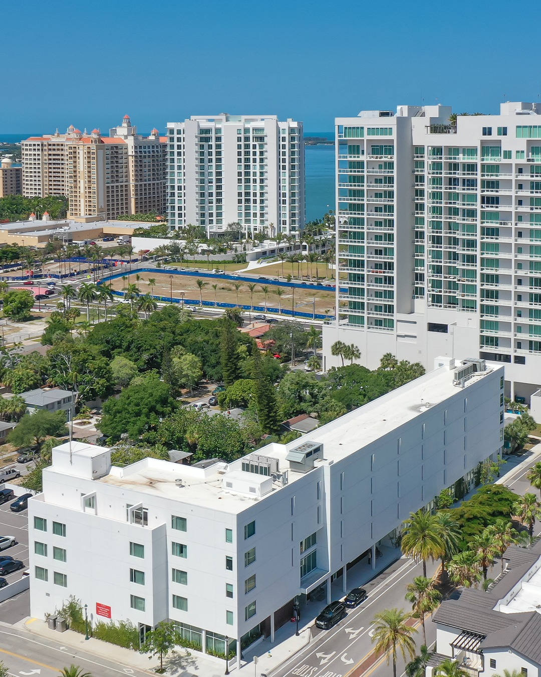 An aerial view of a modern urban area featuring a white multi-story building in the foreground, with taller residential buildings and lush greenery in the background, alongside a glimpse of the ocean in the distance under a clear blue sky.