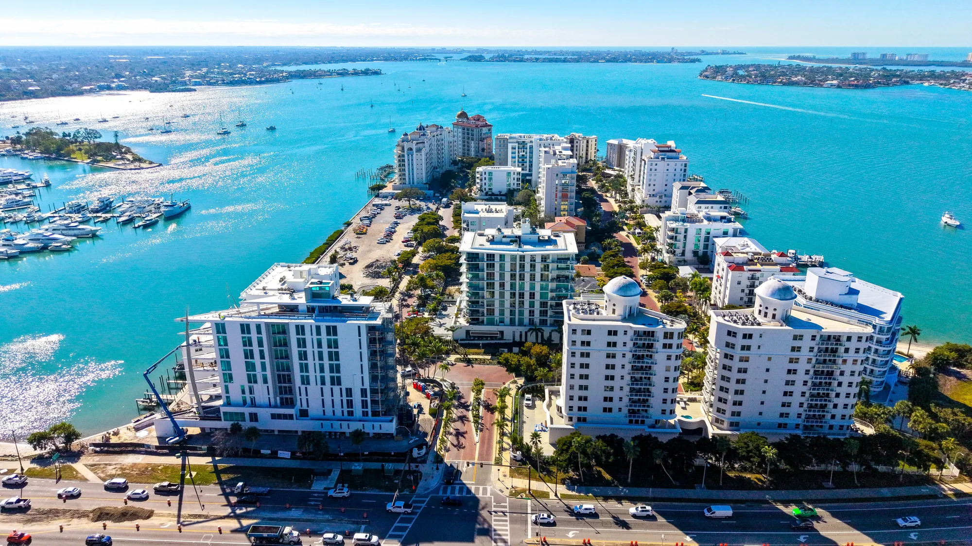 An aerial view of a coastal marina with numerous boats docked, alongside modern residential buildings. The turquoise waters reflect sunlight, creating a scenic backdrop for the developed shoreline.