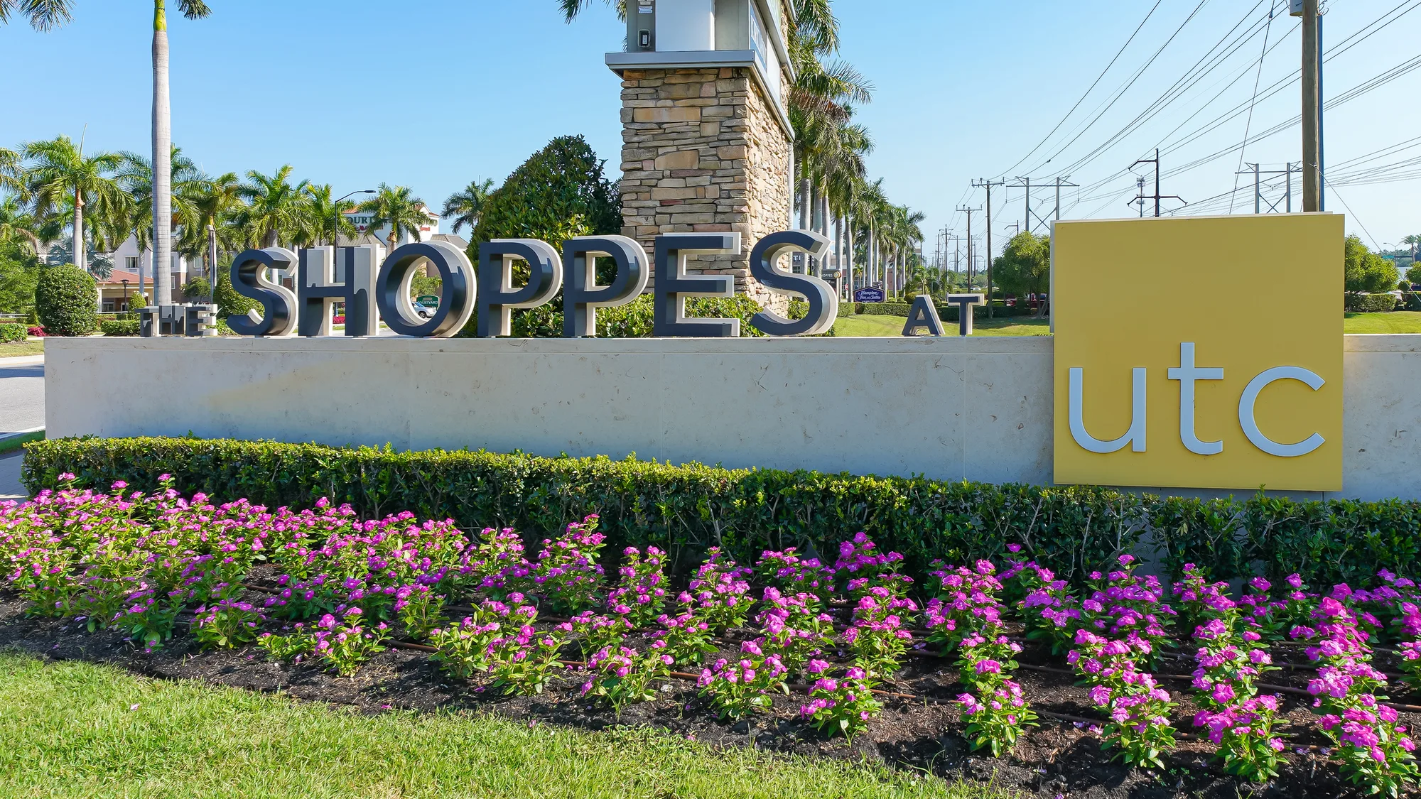 The image depicts a prominent entrance sign for 'The Shoppes at UTC', featuring large letters against a light-colored background. The sign is surrounded by well-maintained landscaping with vibrant purple flowers and palm trees under a clear blue sky.