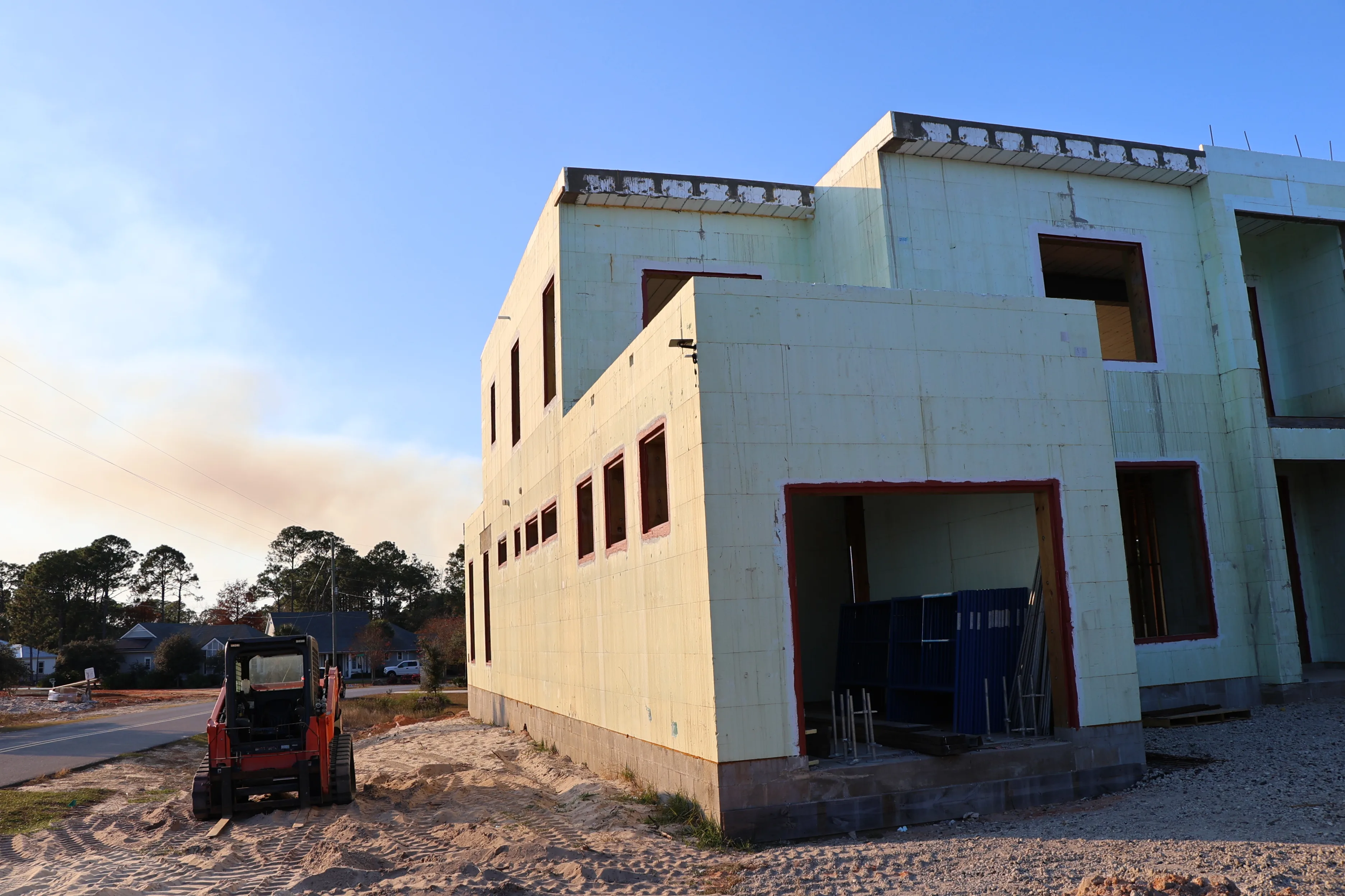 A partially built ICF house beside a small orange excavator on a sandy cleared lot.