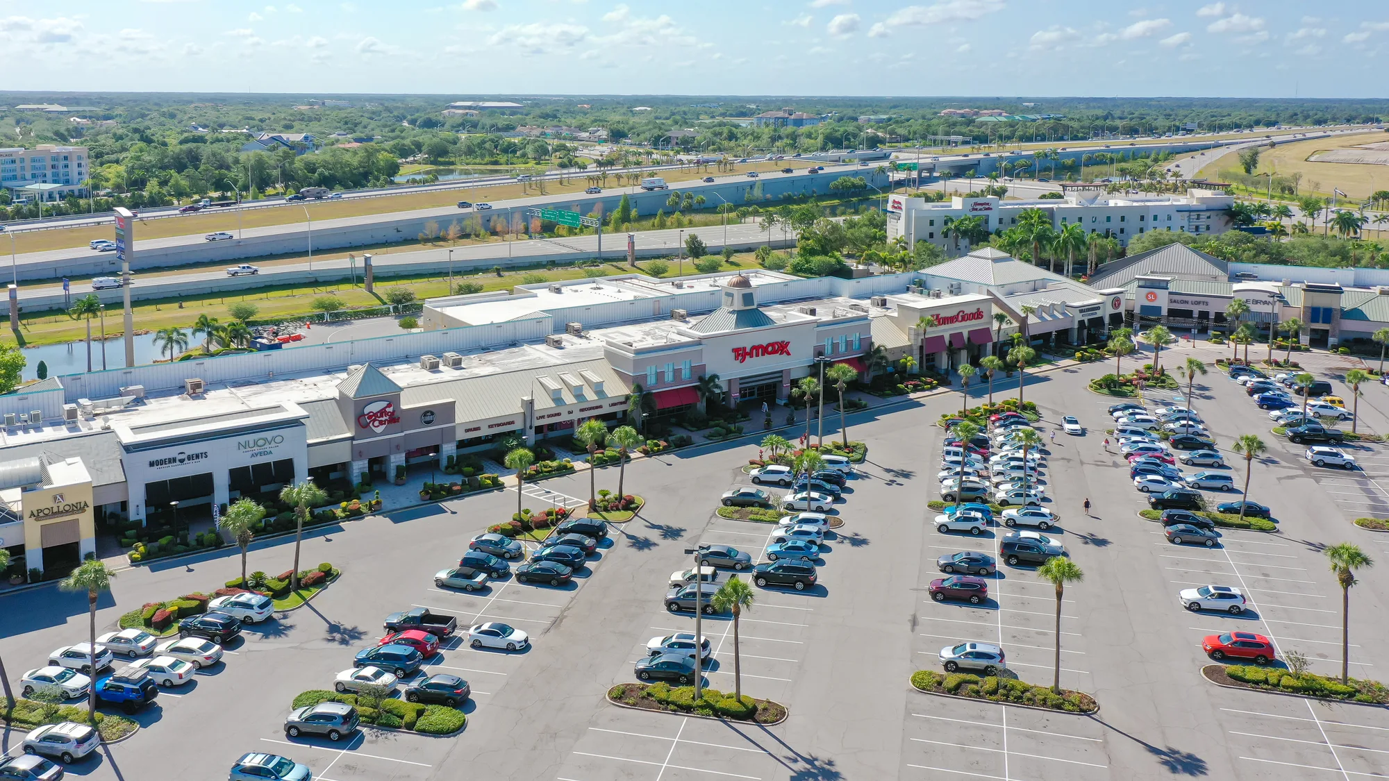 This aerial image shows a busy shopping center with various stores, including TJ Maxx and HomeGoods, surrounded by a large parking lot filled with cars. In the background, a highway runs alongside the center, with greenery visible beyond.