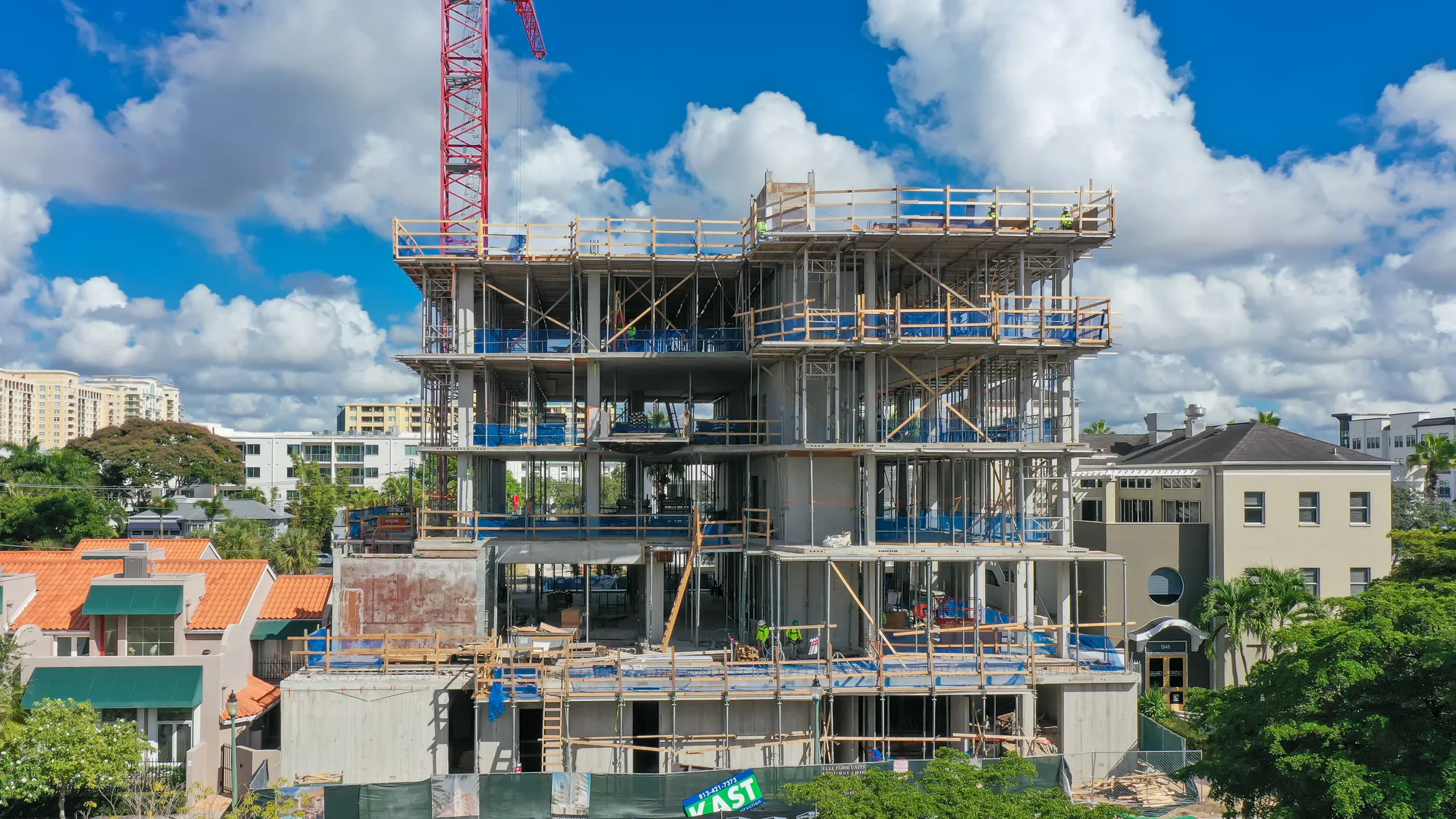 A multi-story building under construction features scaffolding and a crane, set against a backdrop of clouds and neighboring structures. Construction workers can be seen on the site, and the area is surrounded by residential buildings and greenery.