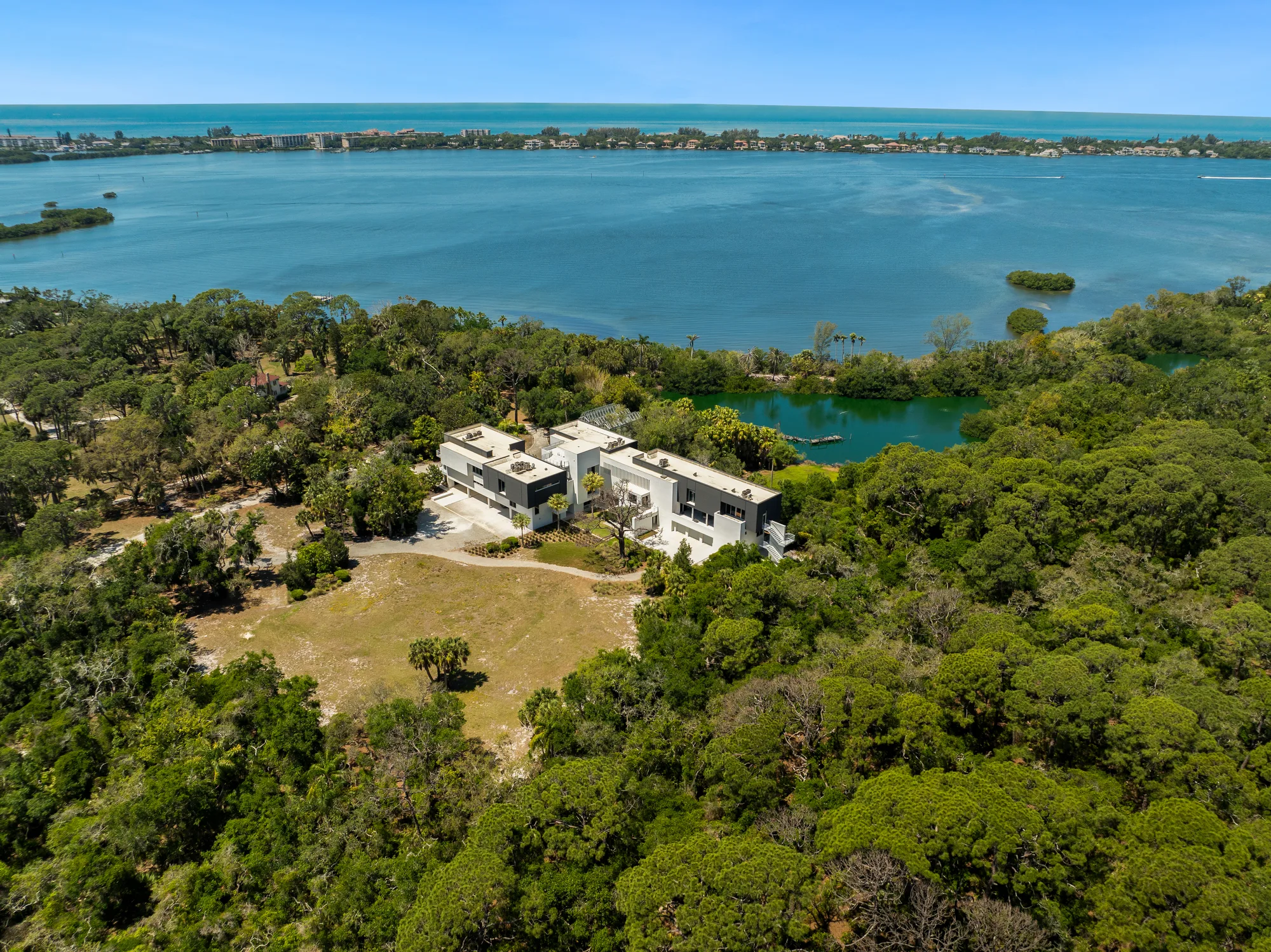 An aerial view of a modern residential house situated near a waterfront, surrounded by lush greenery and trees. The image showcases the home’s expansive outdoor space and proximity to a serene body of water.
