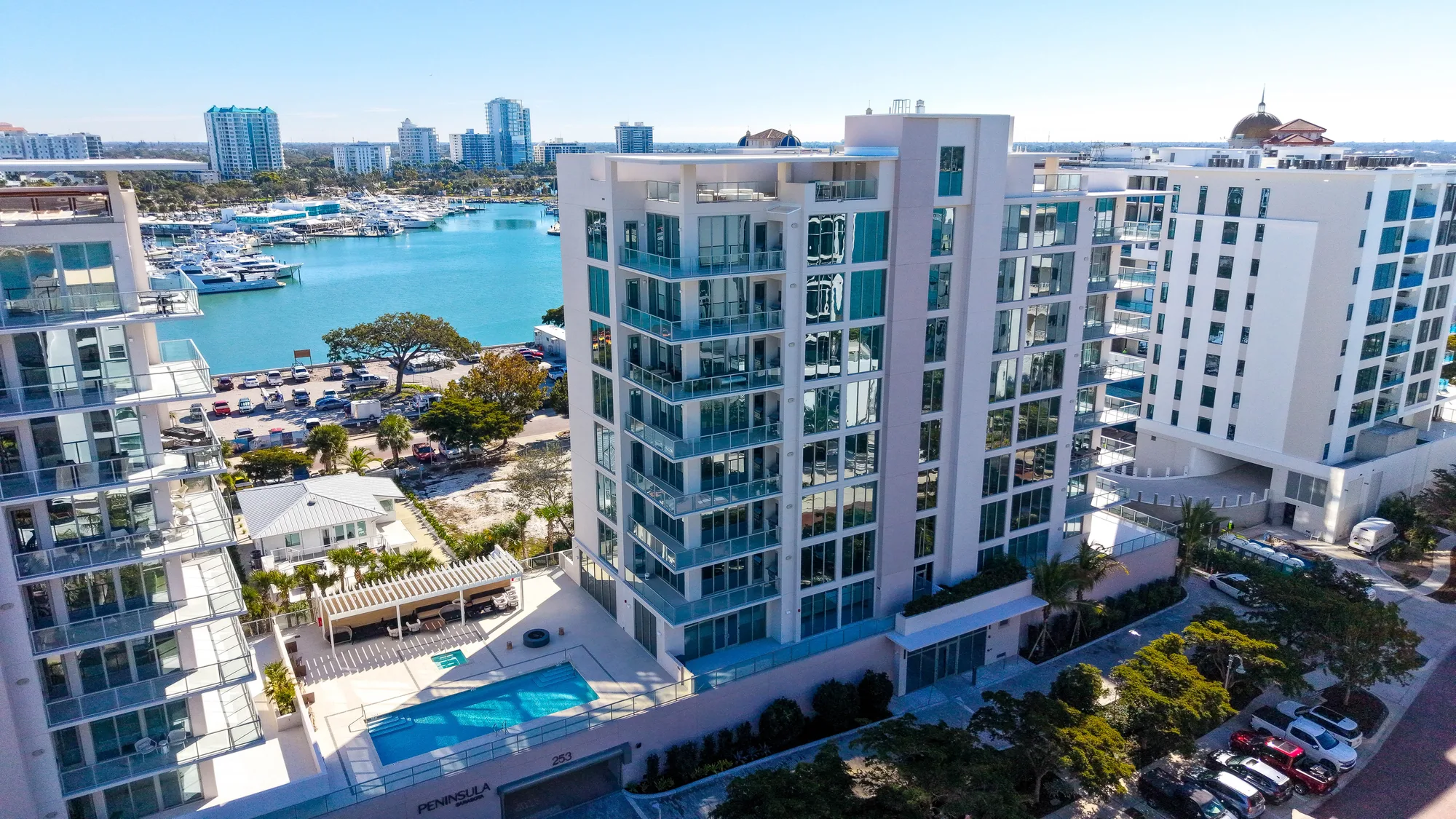 Aerial of the white multi-storey Peninsula Residences building with its pool deck, the Sarasota marina behind it.