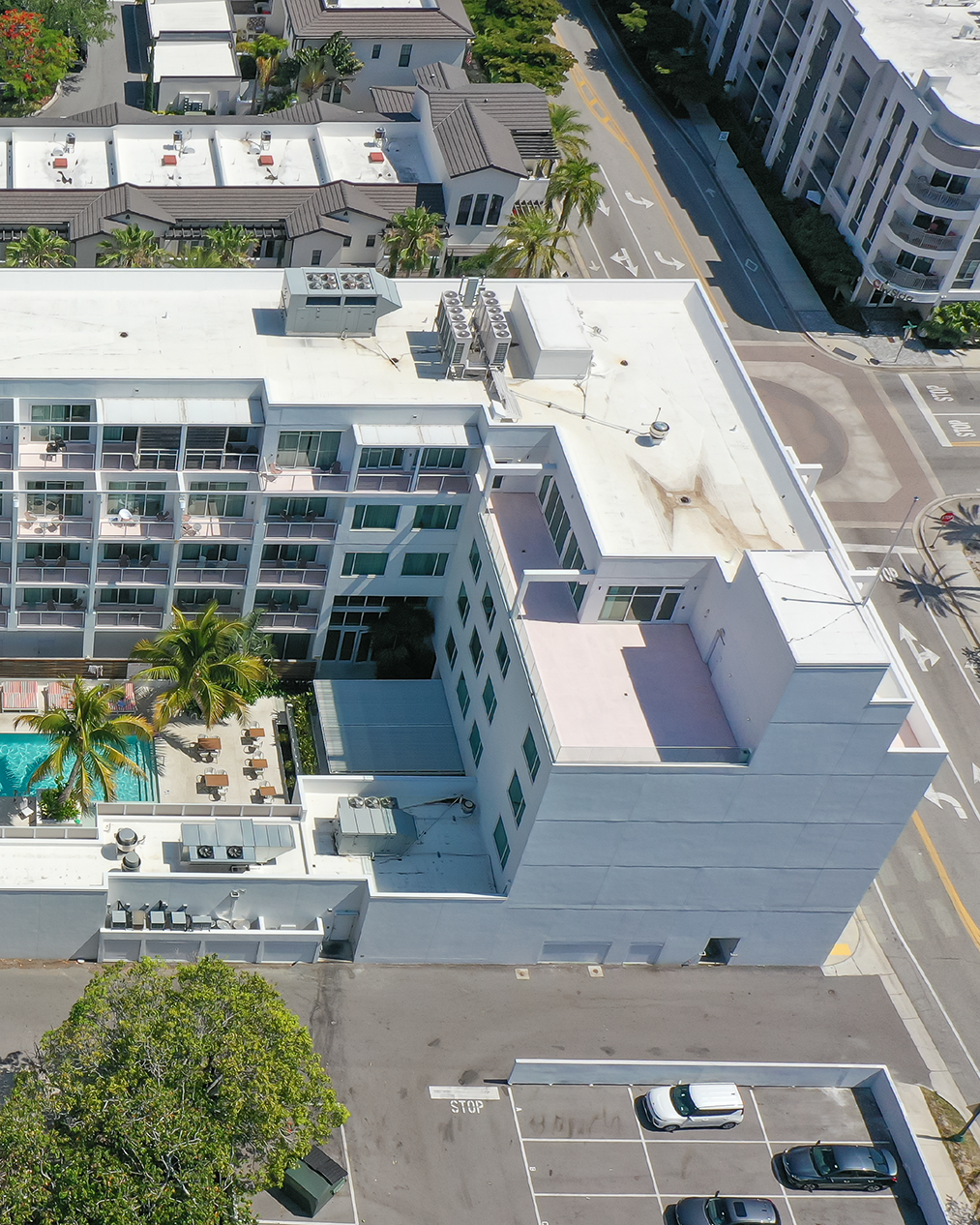 The image depicts an aerial view of a multi-story hotel complex featuring a swimming pool surrounded by lounge chairs. Adjacent buildings and a parking lot are visible, emphasizing the urban setting.