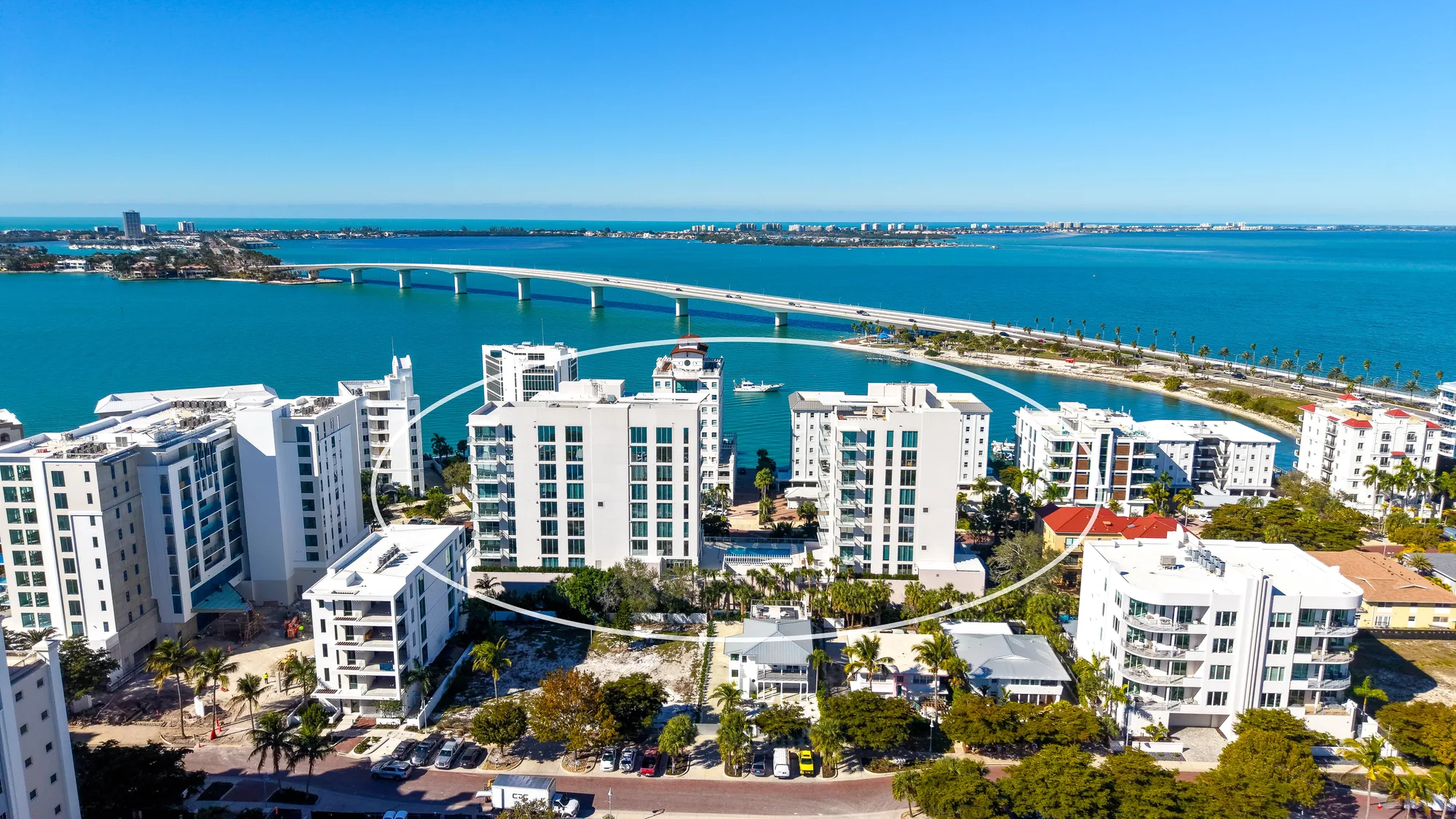 An aerial view of a coastal city featuring modern residential buildings alongside a turquoise bay. A bridge spans the water, connecting the shoreline, with palm trees and a clear blue sky completing the picturesque scene.