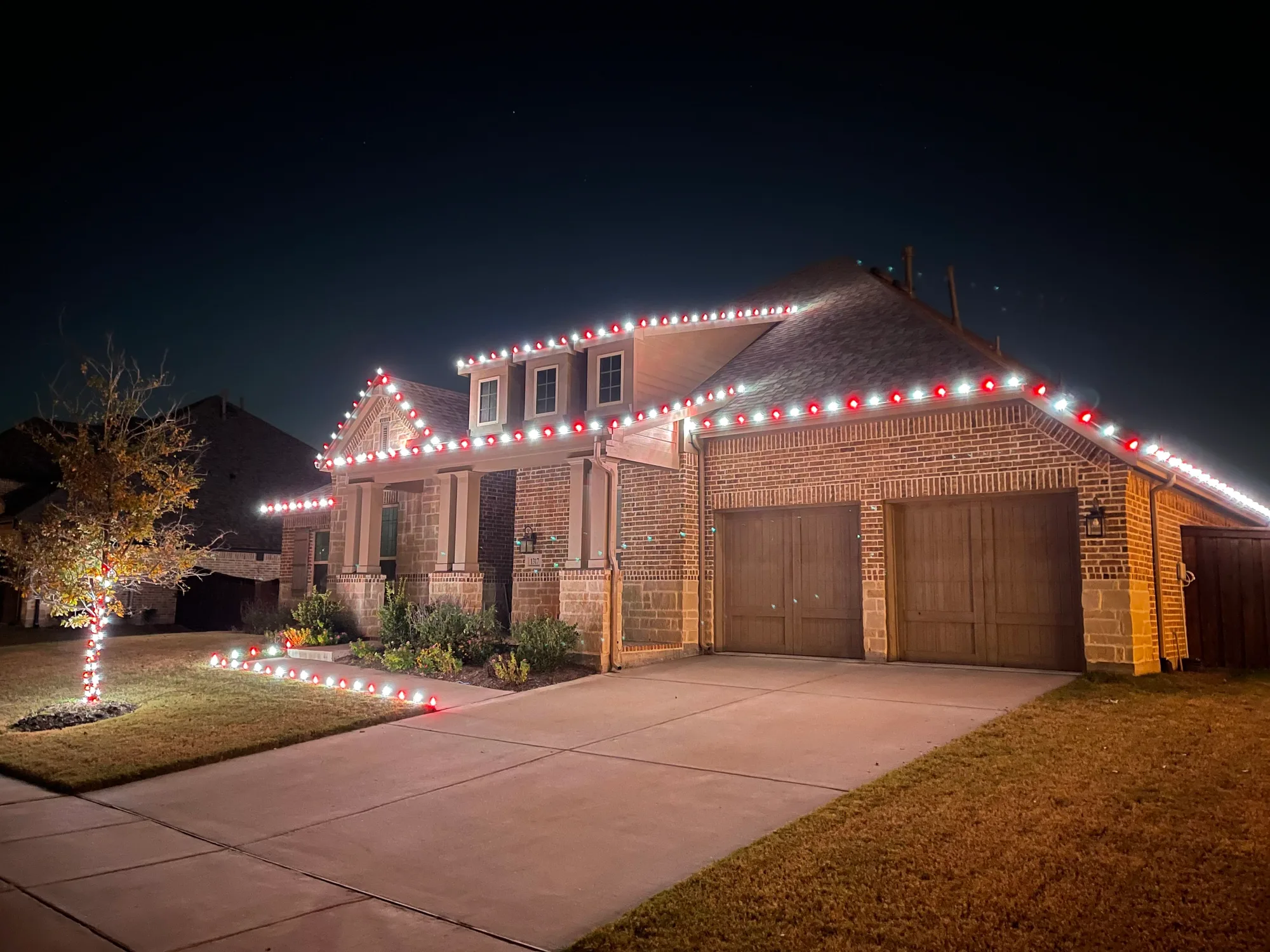 Close view of a roofline showing evenly spaced red and warm white C9 bulbs cut to fit the run, with pathway stake lights and a wrapped tree trunk in the foreground.