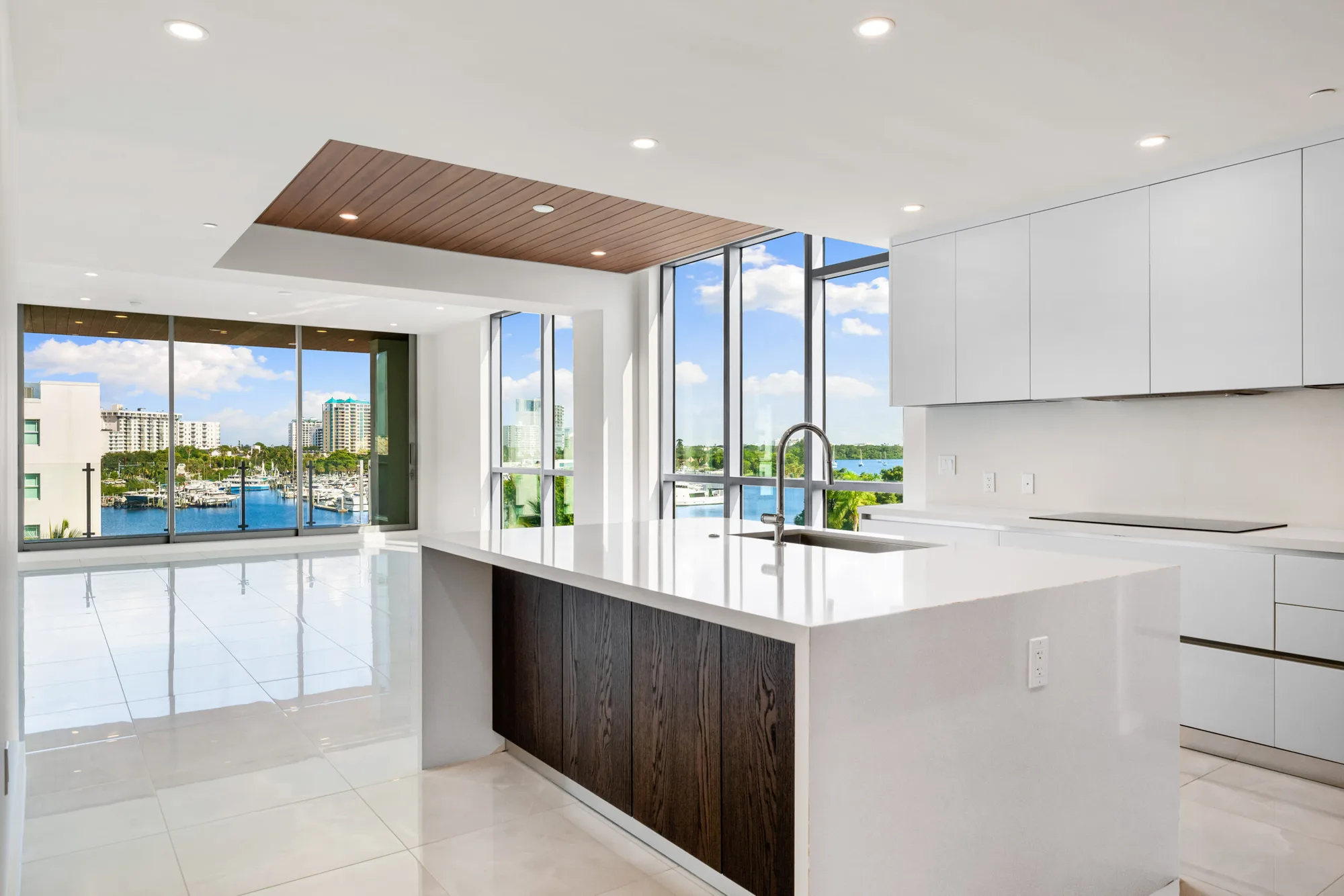 This image showcases a contemporary kitchen featuring a sleek white countertop and cabinetry, complemented by a wooden accent on the island. Large glass windows provide a panoramic view of a marina and urban scenery beyond.