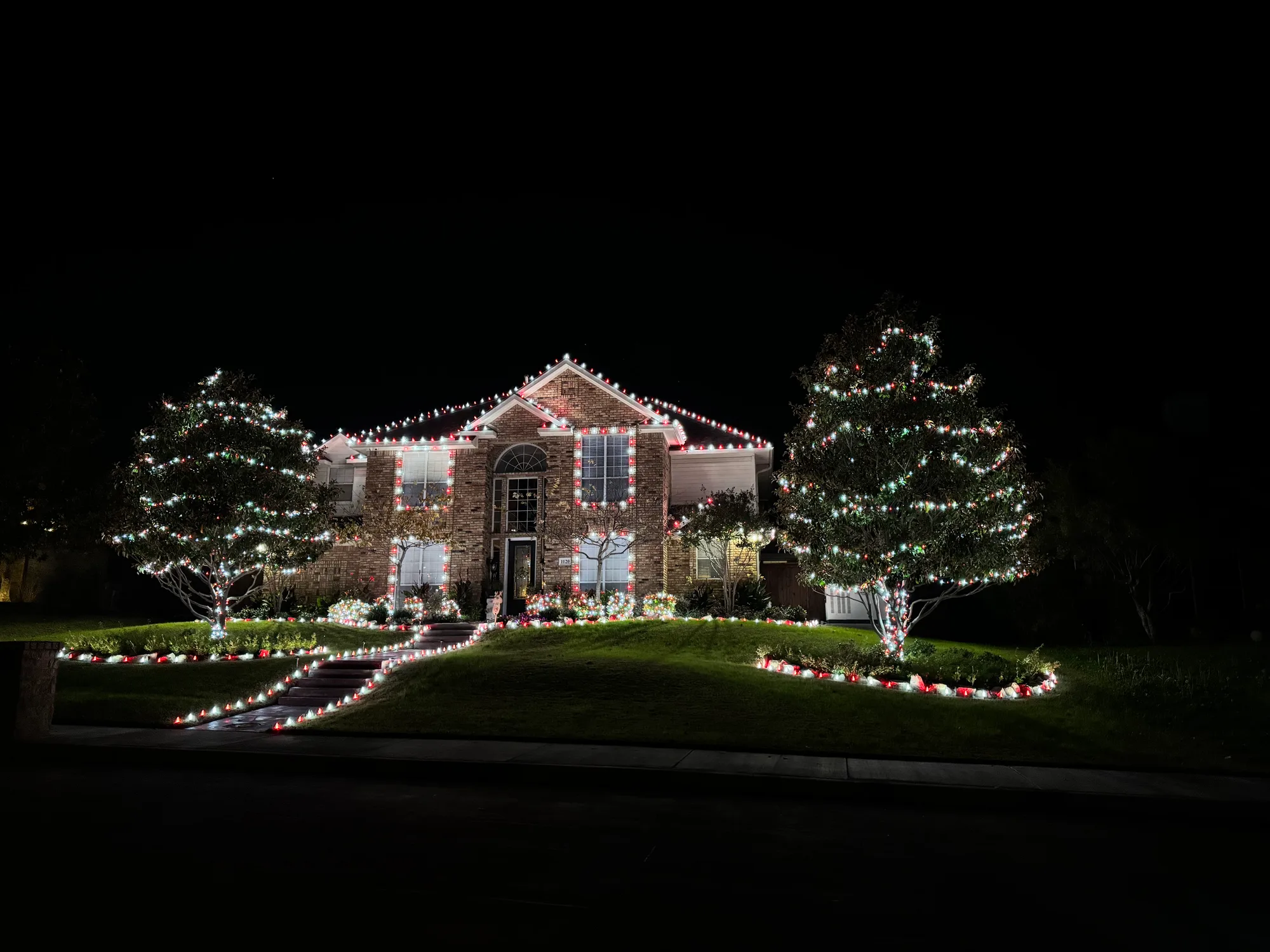 Two-storey home with a red and white roofline and outlined windows, two large wrapped trees, lit garden beds and pathway stake lights along the walkway.