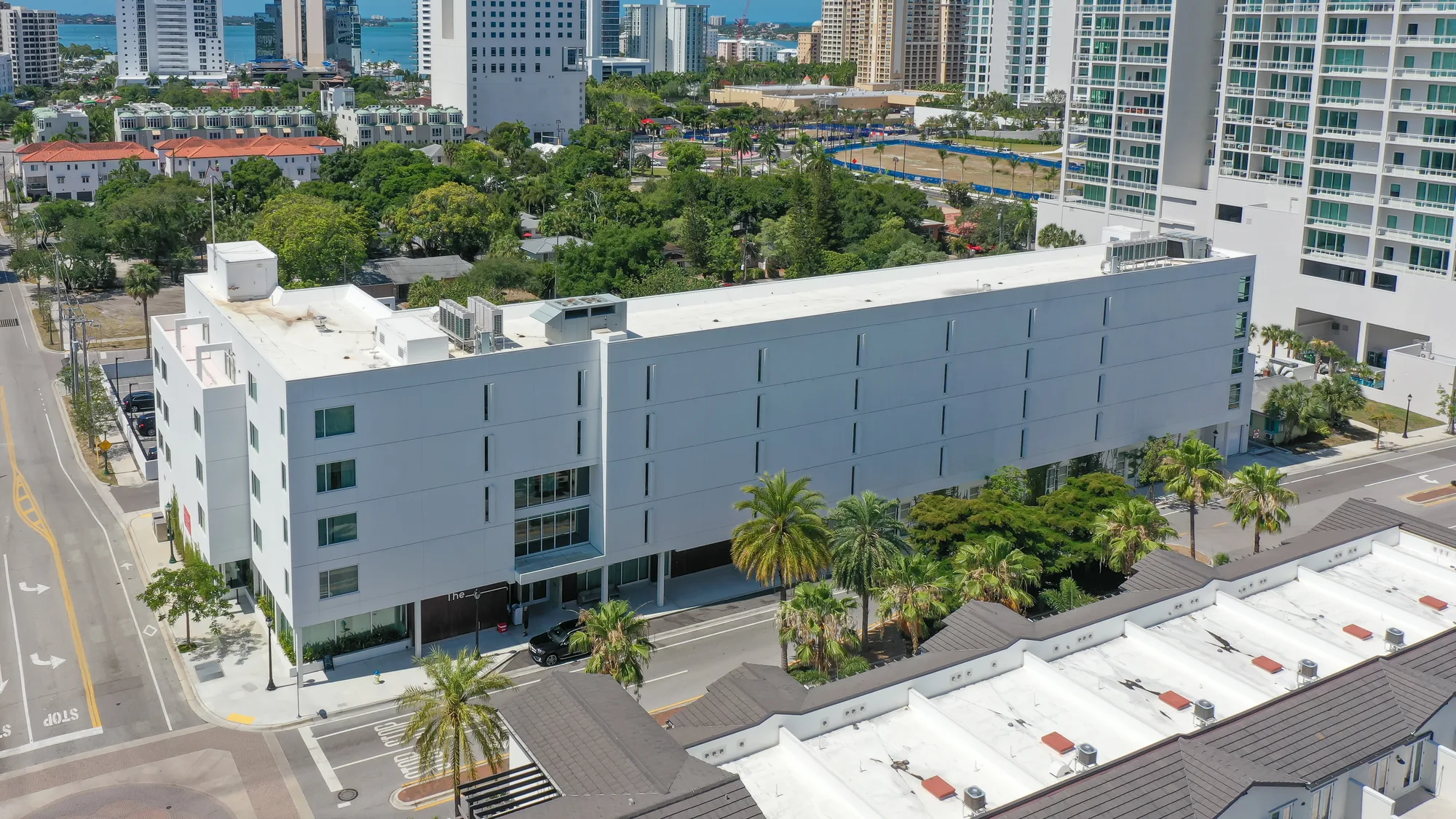 An aerial view of a contemporary white building surrounded by palm trees, with residential homes and high-rise buildings in the background. The image captures a vibrant urban area, showcasing a mix of architecture and greenery.