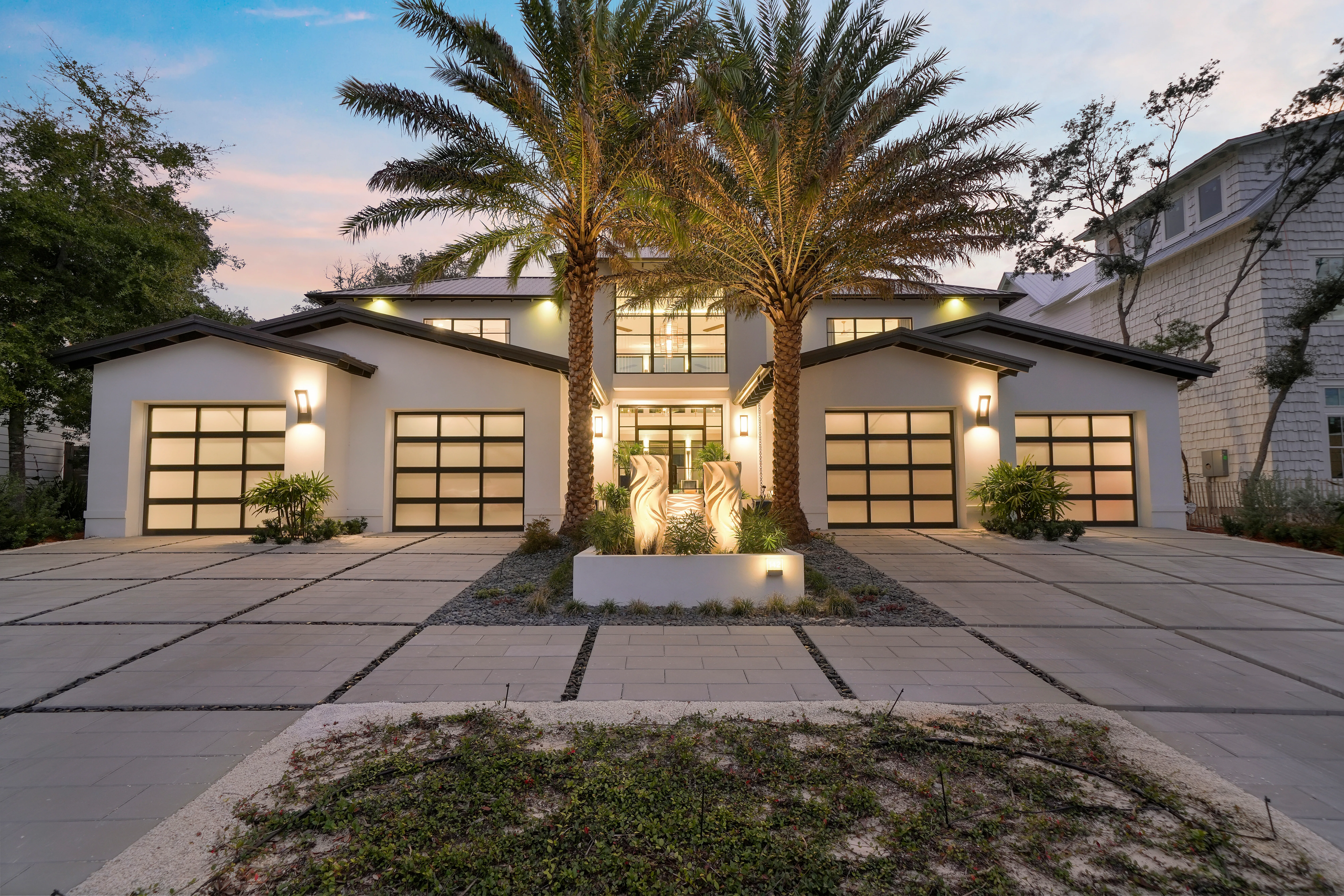 A finished home at night with uplit palm trees over a paver driveway.