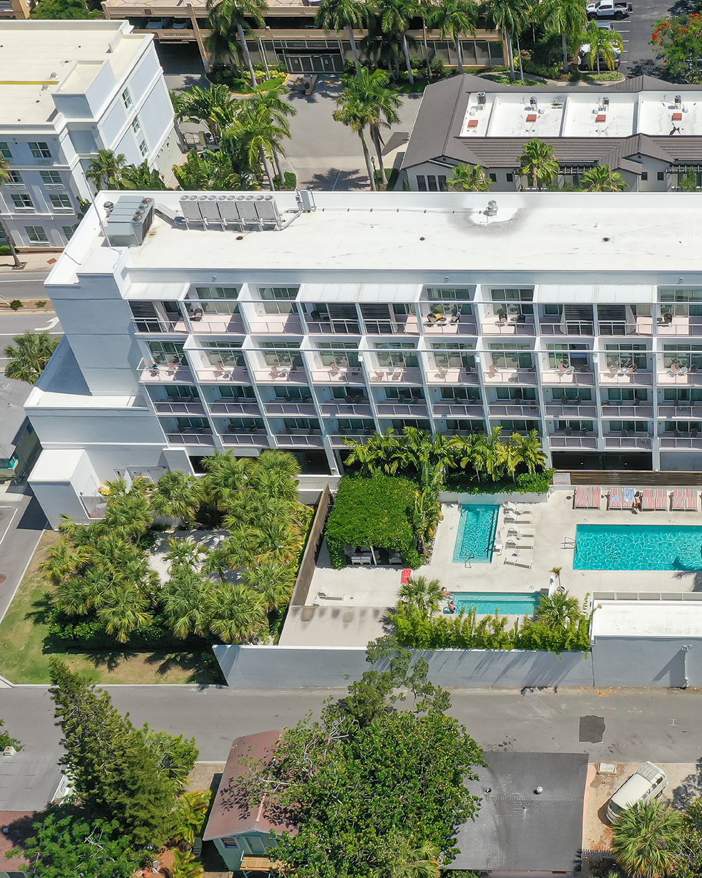 An aerial perspective of a modern hotel featuring multiple floors and balconies, surrounded by lush greenery. The image shows a swimming pool area with lounge chairs and palms, highlighting the tranquil setting.