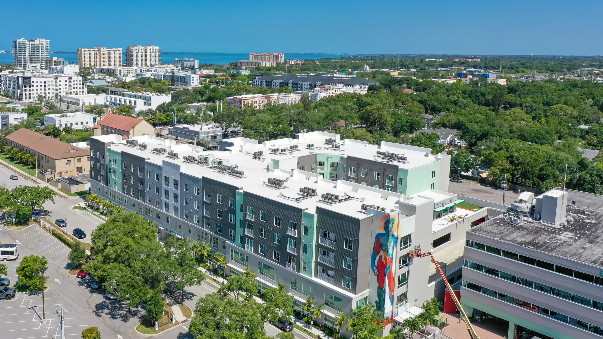 An aerial view of a contemporary apartment building surrounded by greenery and urban development. The building features colorful murals and is located near a coastal area, with additional apartment complexes and cityscape visible in the background.