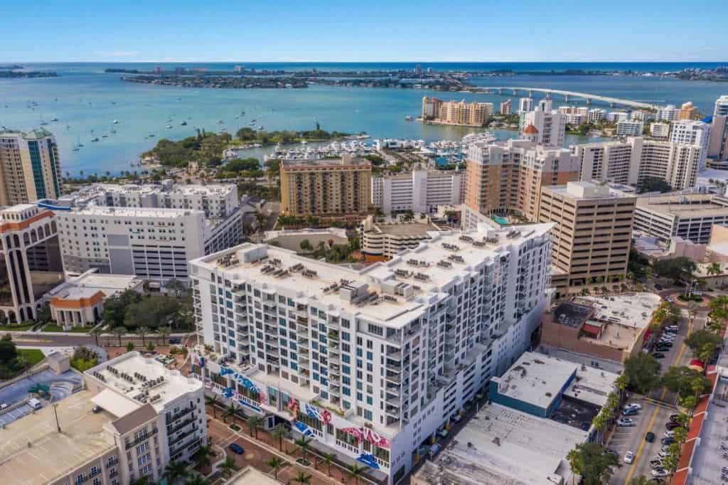 The image showcases an aerial view of a coastal urban area featuring modern buildings and lush greenery, with a serene bay in the background. Boats can be seen on the water, and the skyline includes a mix of residential and commercial structures.