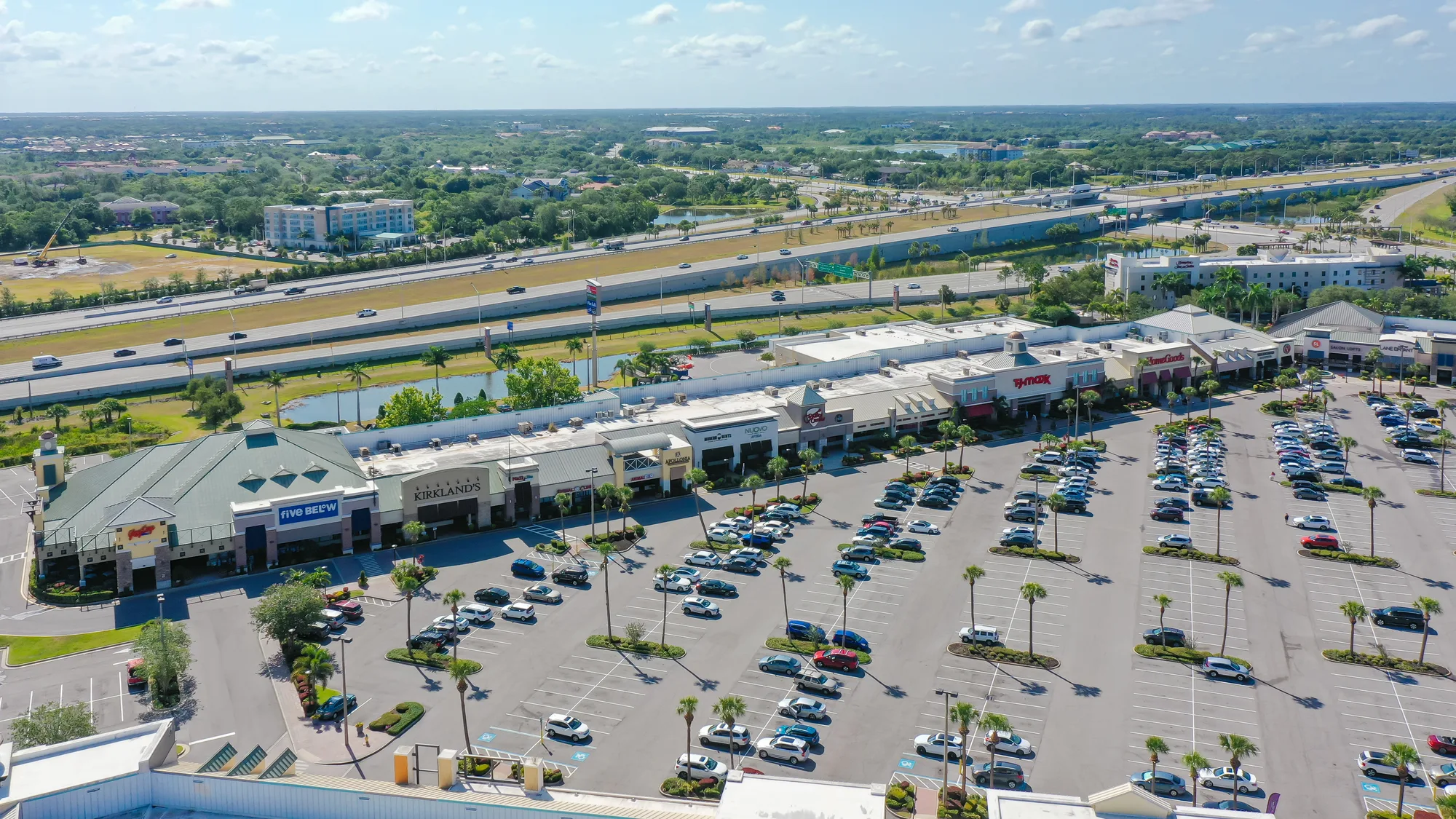 This image shows an aerial view of a shopping center with a wide parking lot filled with cars. The surrounding area includes a highway and green landscape, with several retail stores visible along the front.
