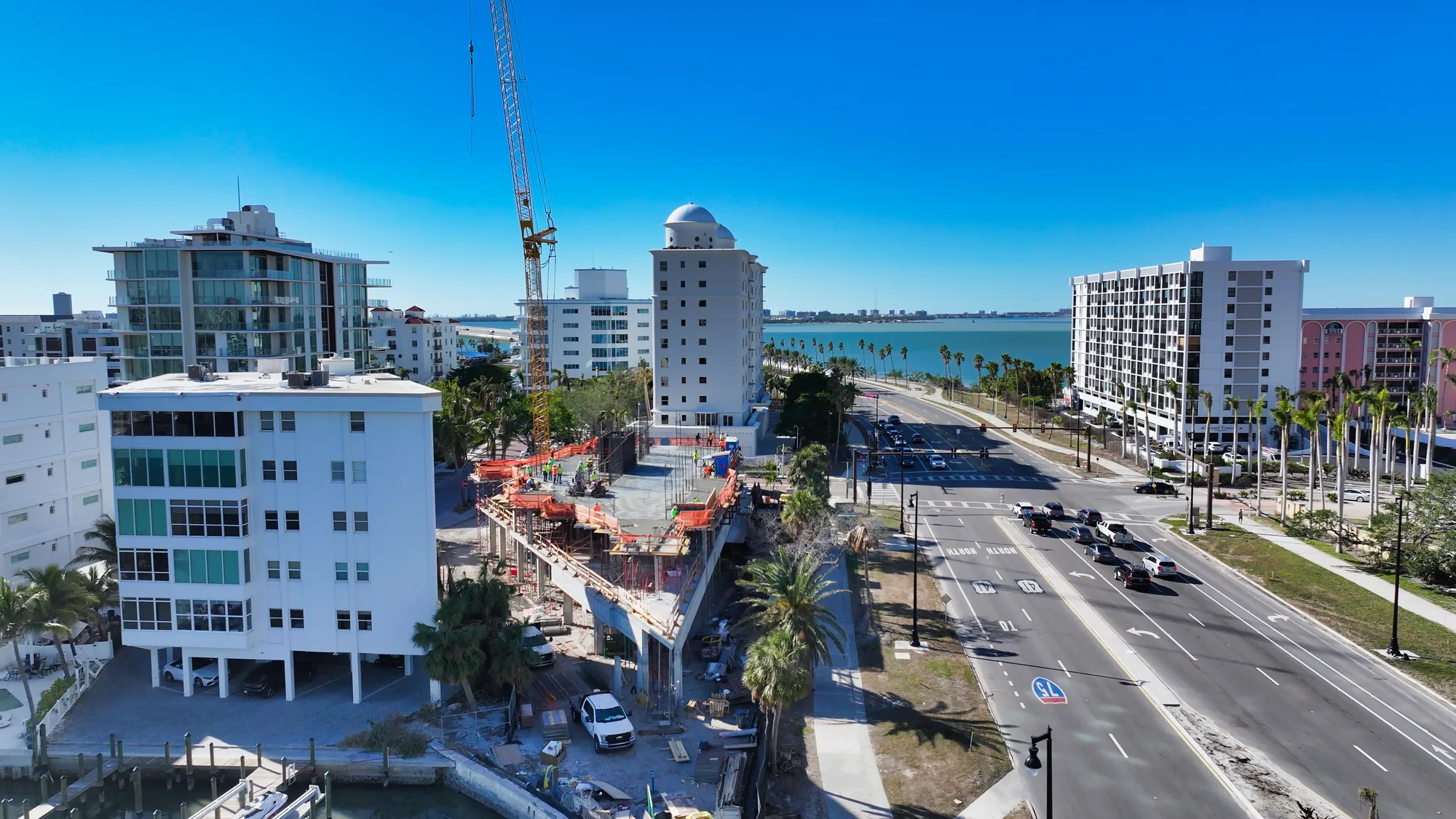This image captures a coastal construction site with new buildings and a crane, alongside a busy road lined with palm trees and vehicles. The blue ocean and clear sky create a vibrant backdrop for the urban development.