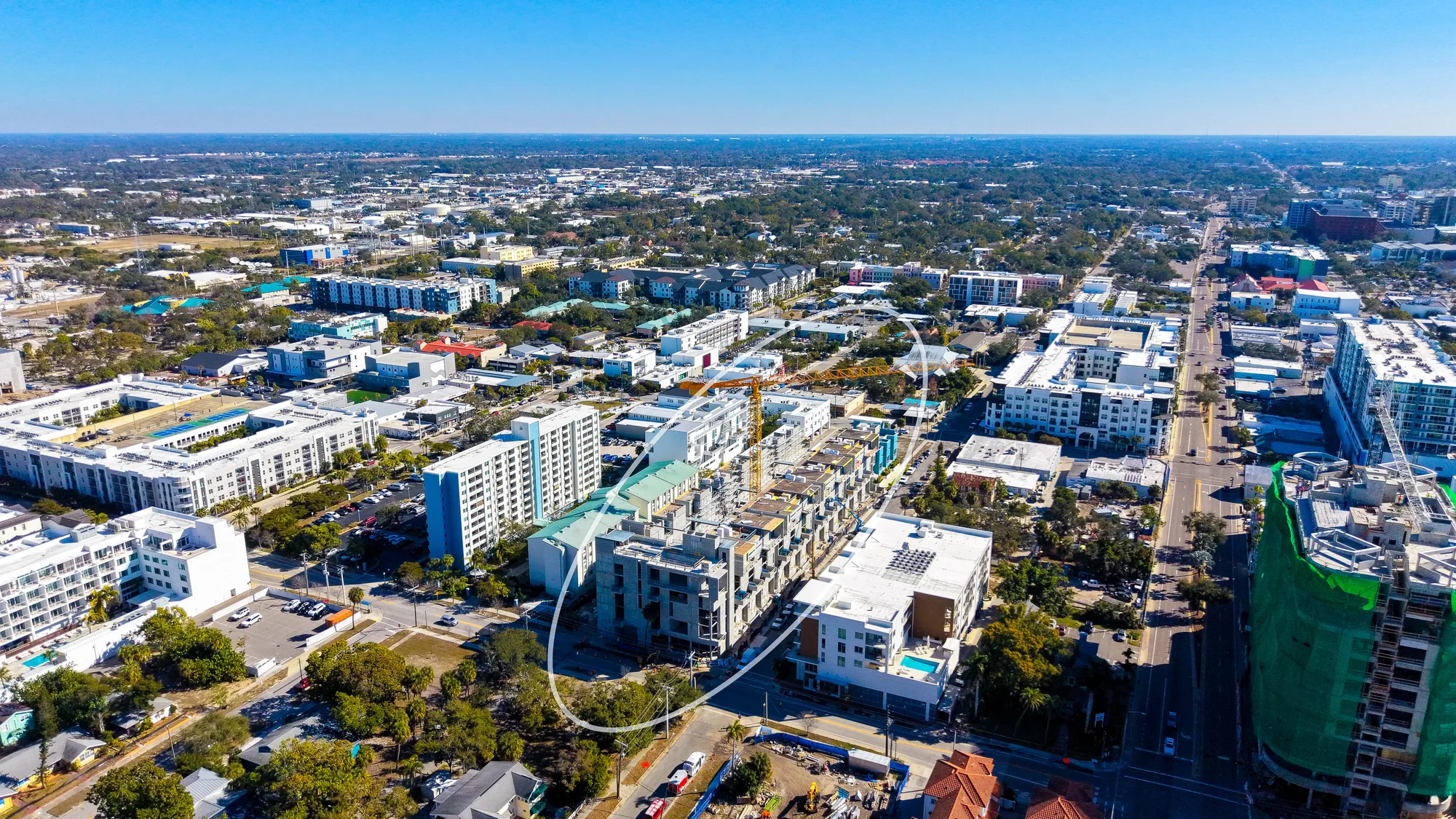The image showcases an aerial perspective of a vibrant urban area featuring various buildings, residential complexes, and ongoing construction. The landscape includes a mix of modern architecture and greenery, set against a clear blue sky.